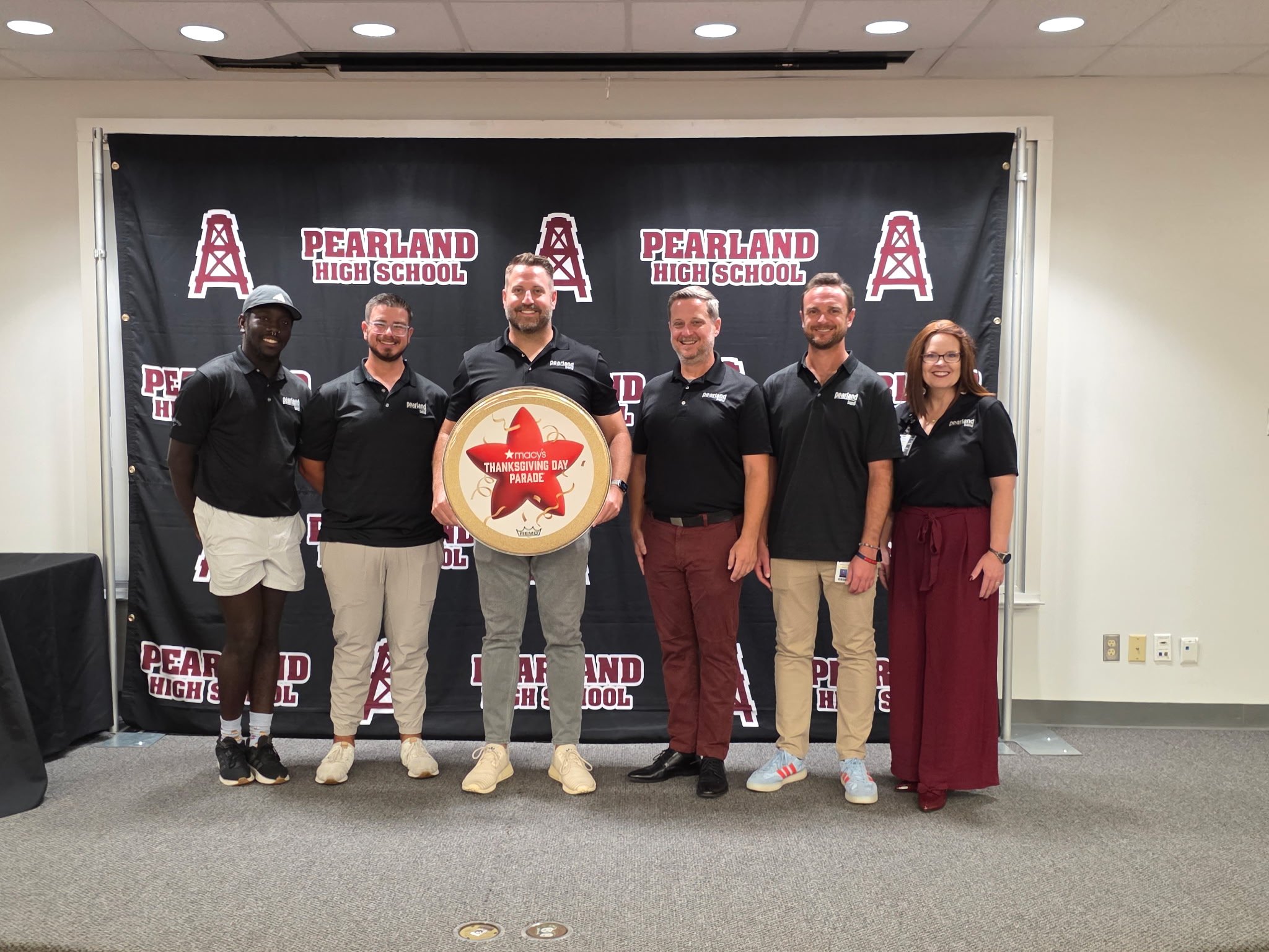 A group of six people standing in front of a black backdrop with 'Pearland High School' logos and red oil rig icons, some holding a large wooden plaque that says 'Macy's Thanksgiving Day Parade'.