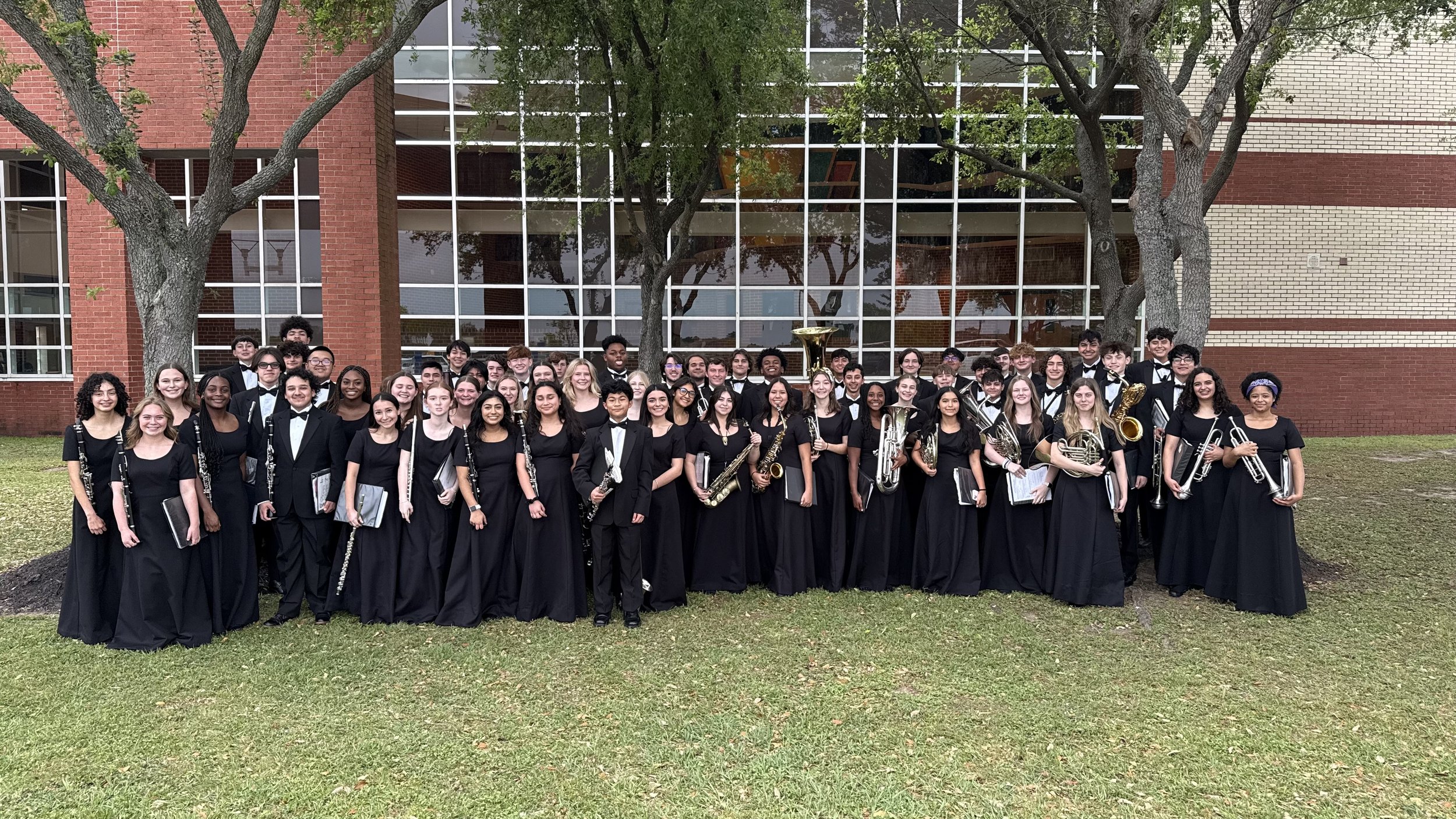 Large group of young musicians in black formal attire, holding musical instruments, posing outside in front of a brick and glass building with trees.