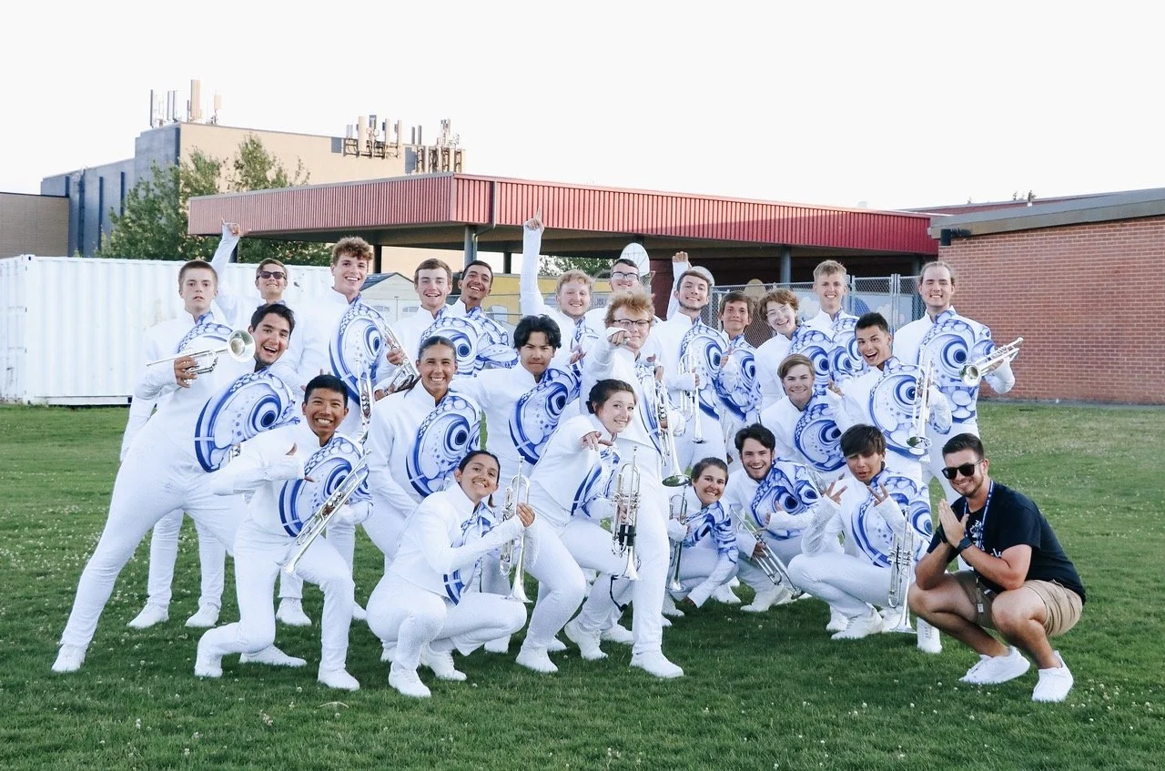 A group of young people in white uniforms with blue spirals holding brass instruments, posing outdoors on a grassy field with a brick building in the background.
