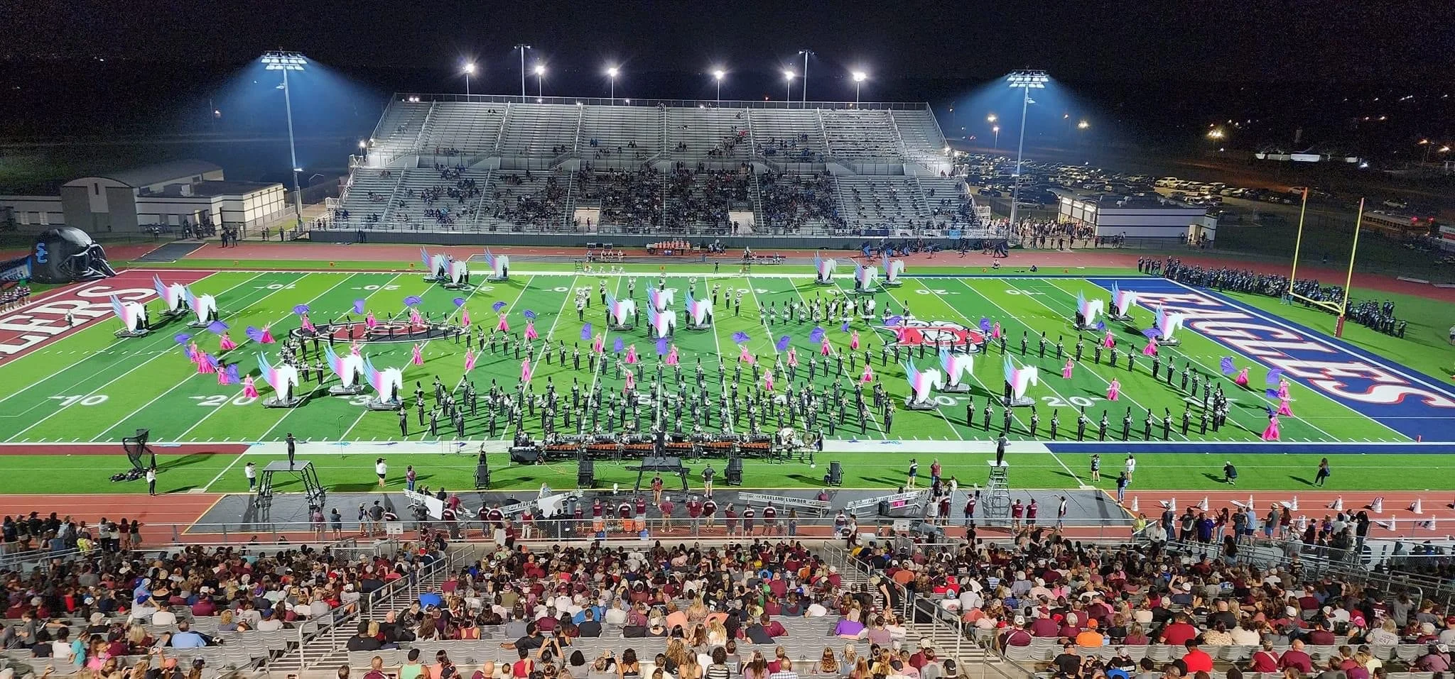 Nighttime scene of a stadium filled with spectators watching a marching band performance on the football field, decorated with colorful banners and props, with bright stadium lights illuminating the field.