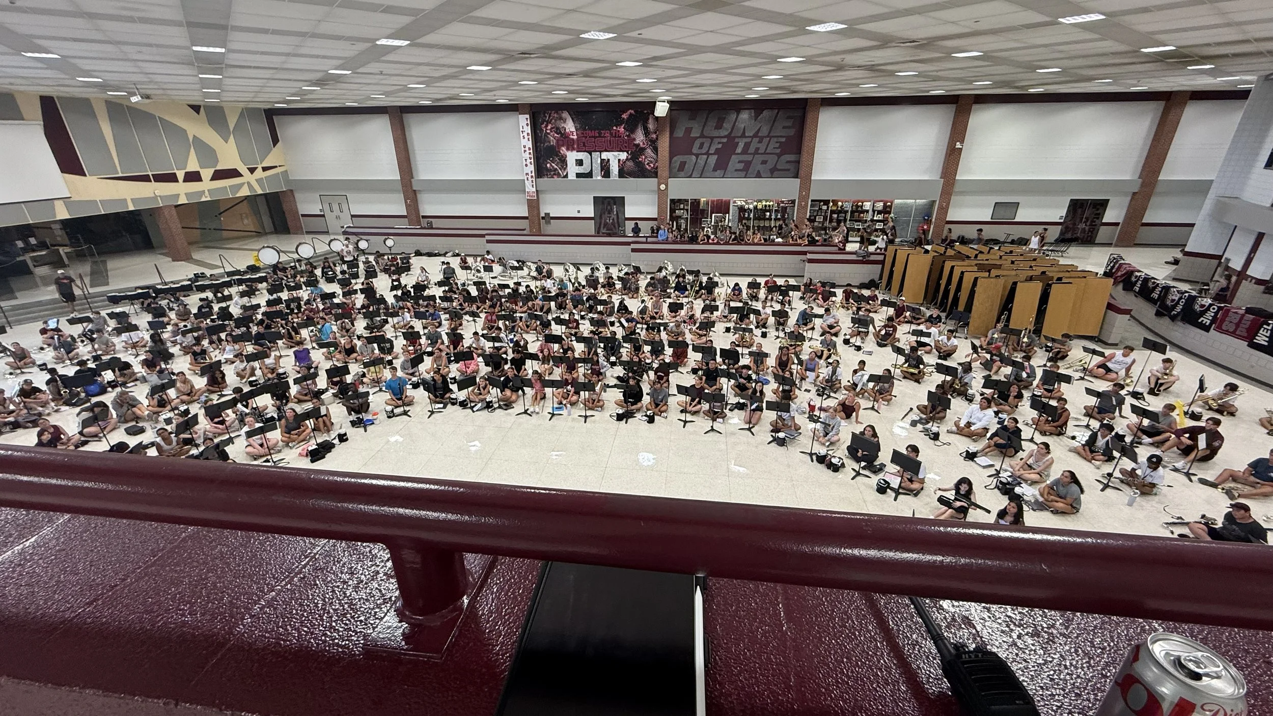 A large indoor auditorium filled with students sitting on the floor in front of music stands, practicing with instrument amplifiers and music equipment. The background features banners and a stage. The ceiling has a grid of fluorescent lights.