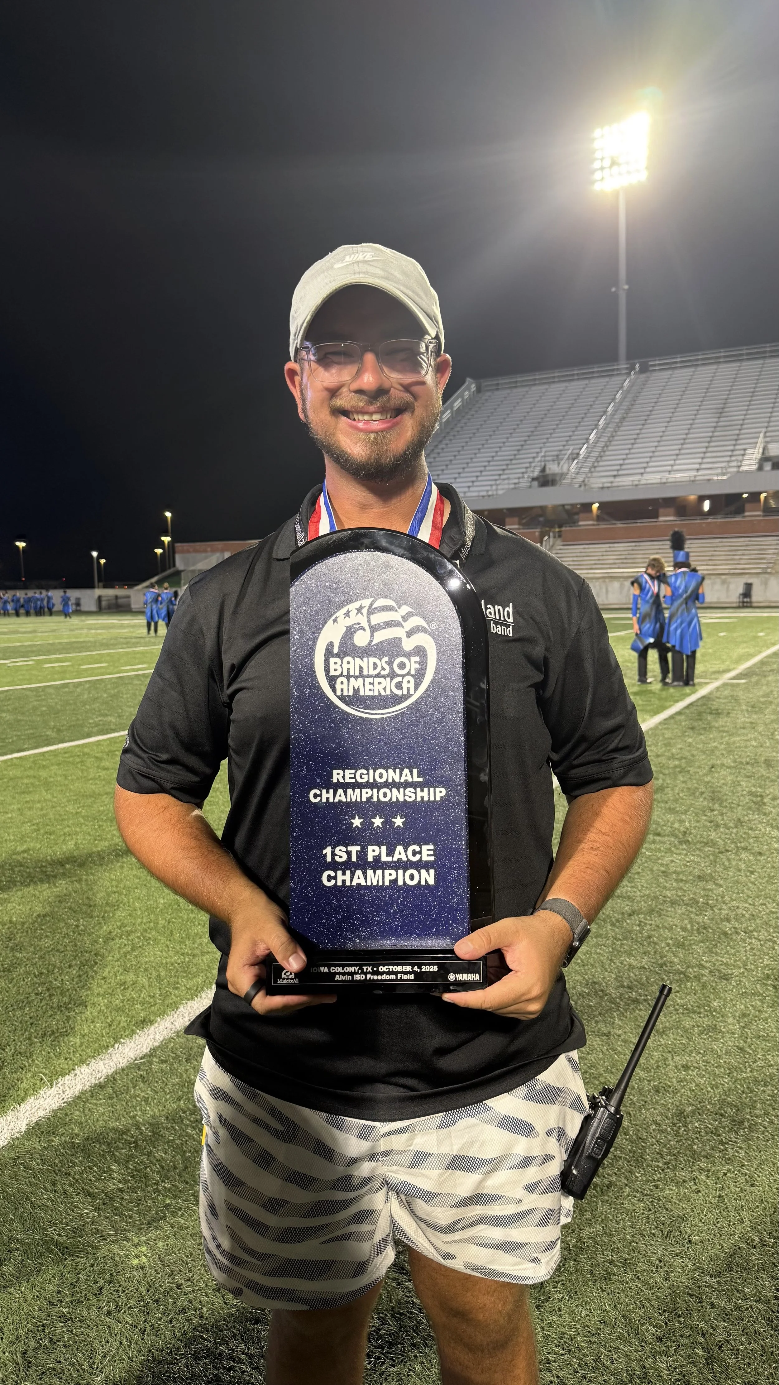 A man holding a trophy on a football field at night, celebrating a first-place win at the Bands of America Regional Championship. He is smiling, wearing glasses, a cap, and athletic clothing, with other people in the background.