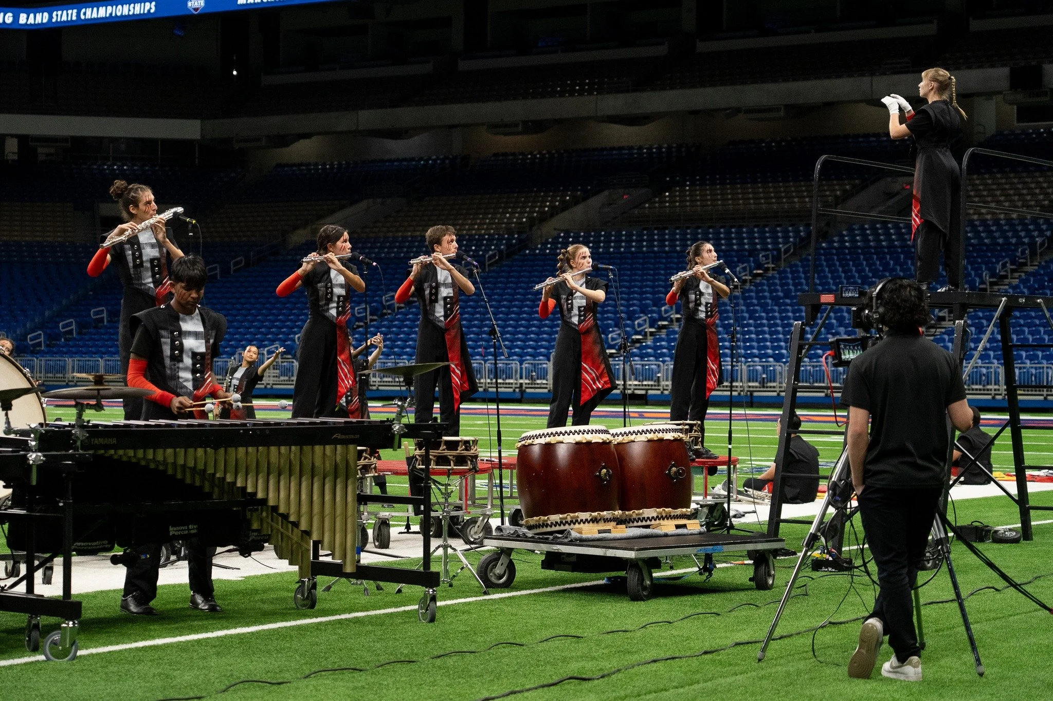 A marching band practice on a football field, with musicians playing wind instruments and percussion, a conductor standing on a platform, and a camera operator recording the scene.