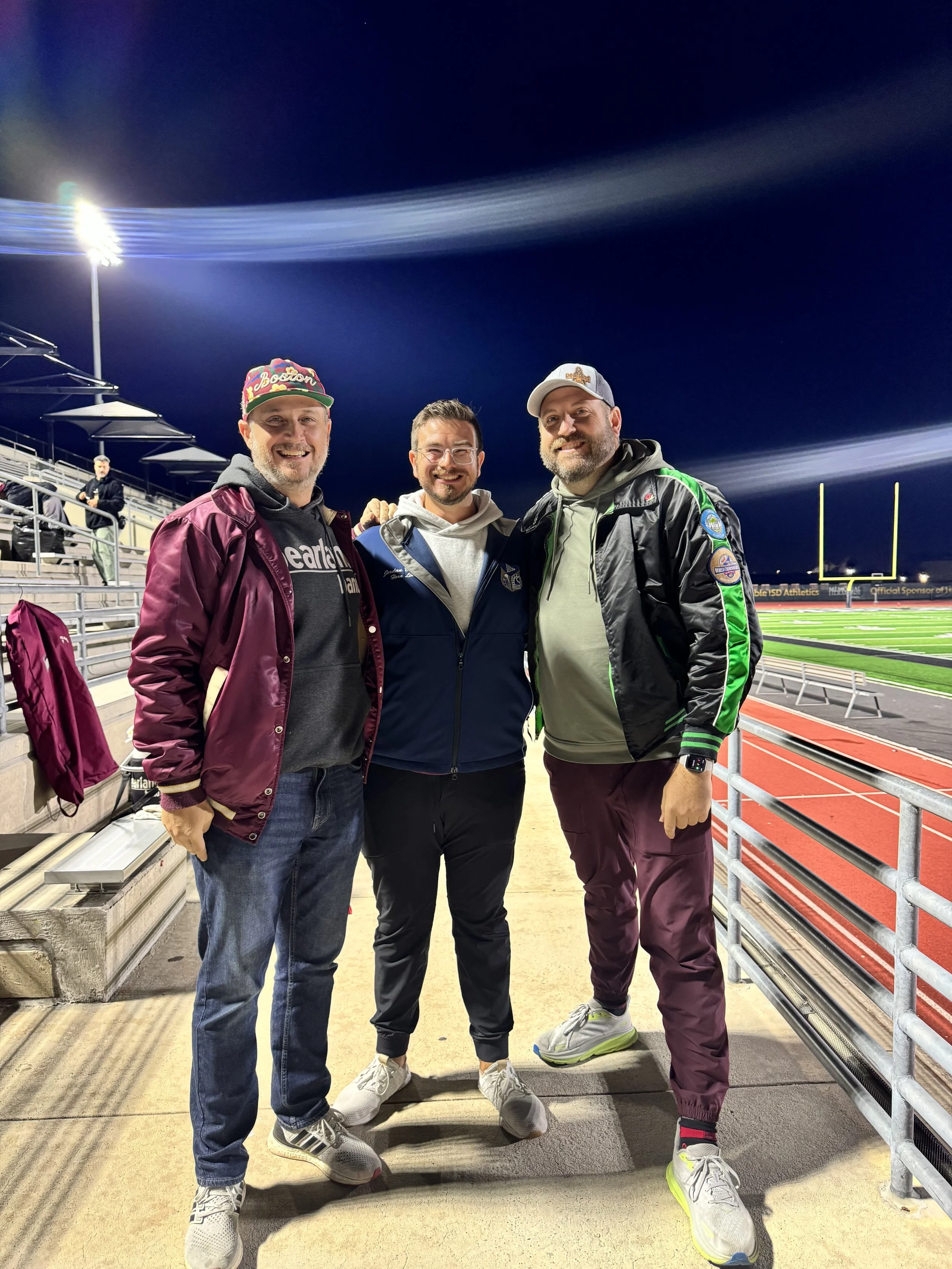 Three men standing together smiling at a stadium at night. The stadium is lit with bright lights, with empty bleachers in the background and a football field behind a railing.