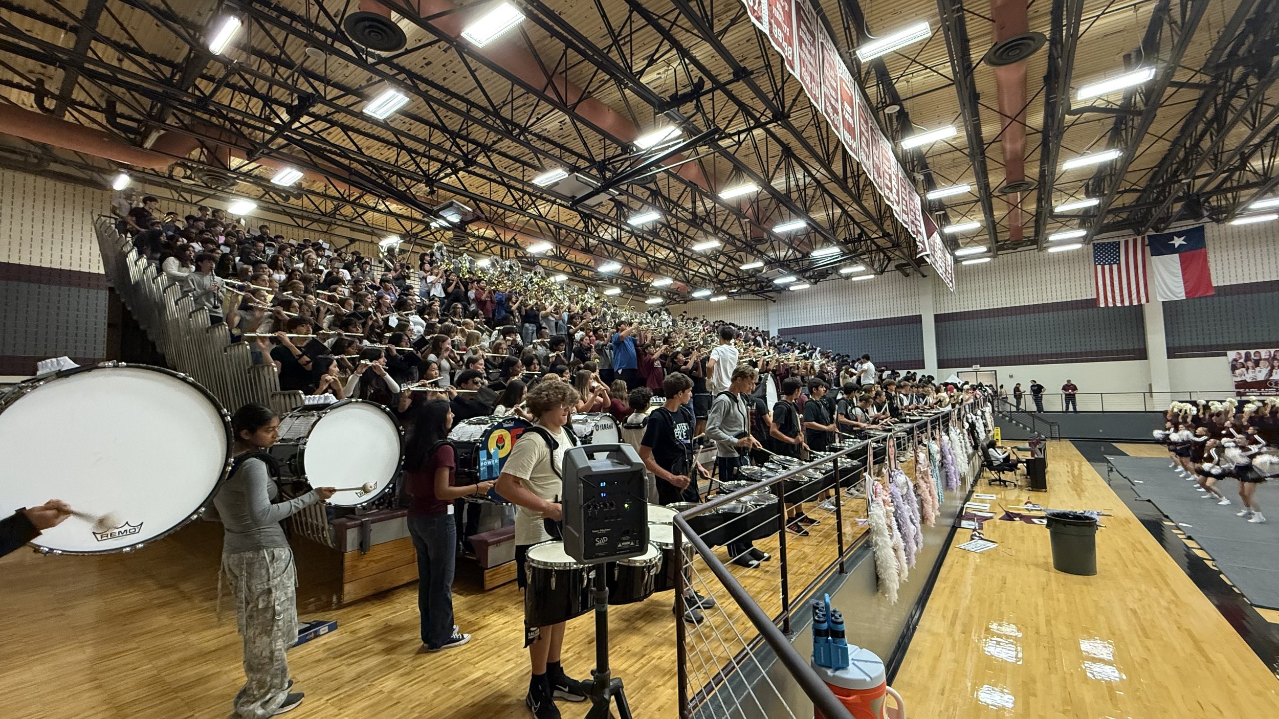 A large indoor gymnasium filled with a school marching band on bleachers, students playing drums, and dancers on the stage, with flags hanging on the wall.