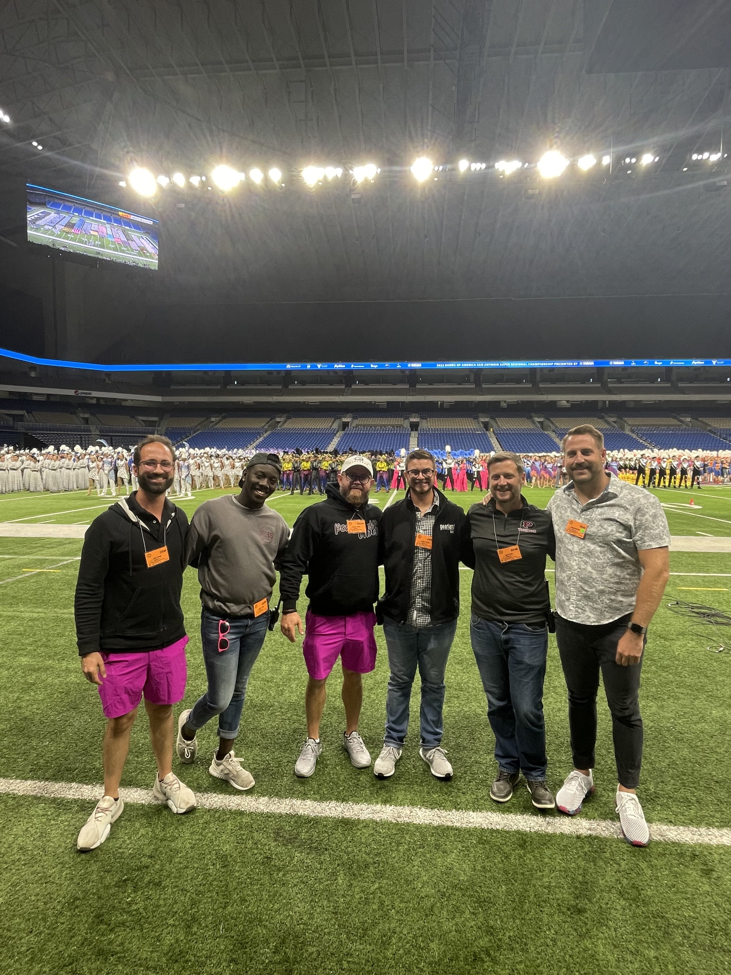 Group of six men standing together on a football field inside a stadium with a large crowd and band in the background.