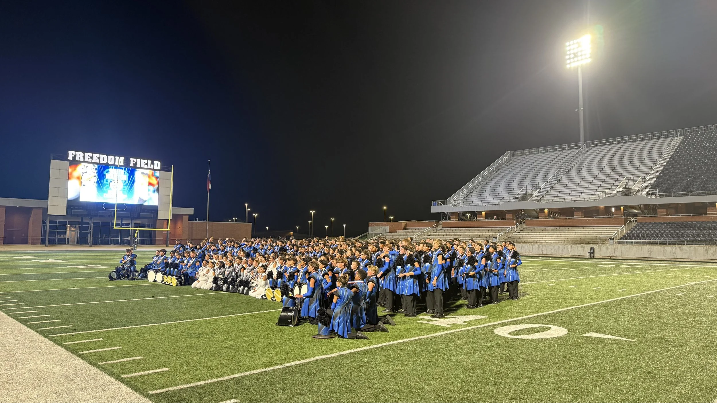 Large group of marching band members in blue and black uniforms kneeling and standing on football field at night, with empty stadium stands and a large screen displaying a graphic and the words "Freedom Field" in the background.