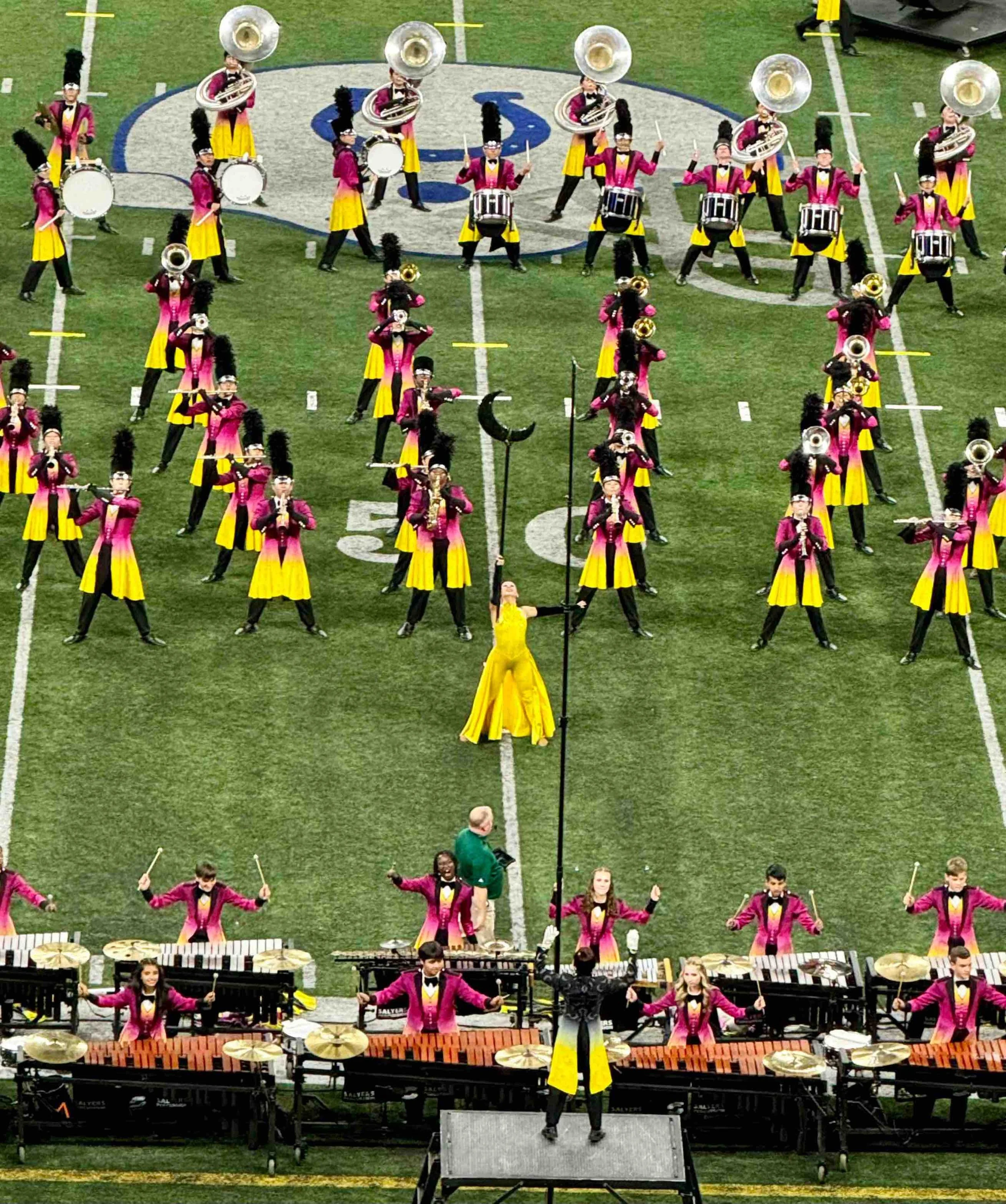 A marching band performing on a football field, wearing magenta and yellow uniforms. The band includes percussion, brass, and woodwind players, with a drum major in a yellow gown leading them.