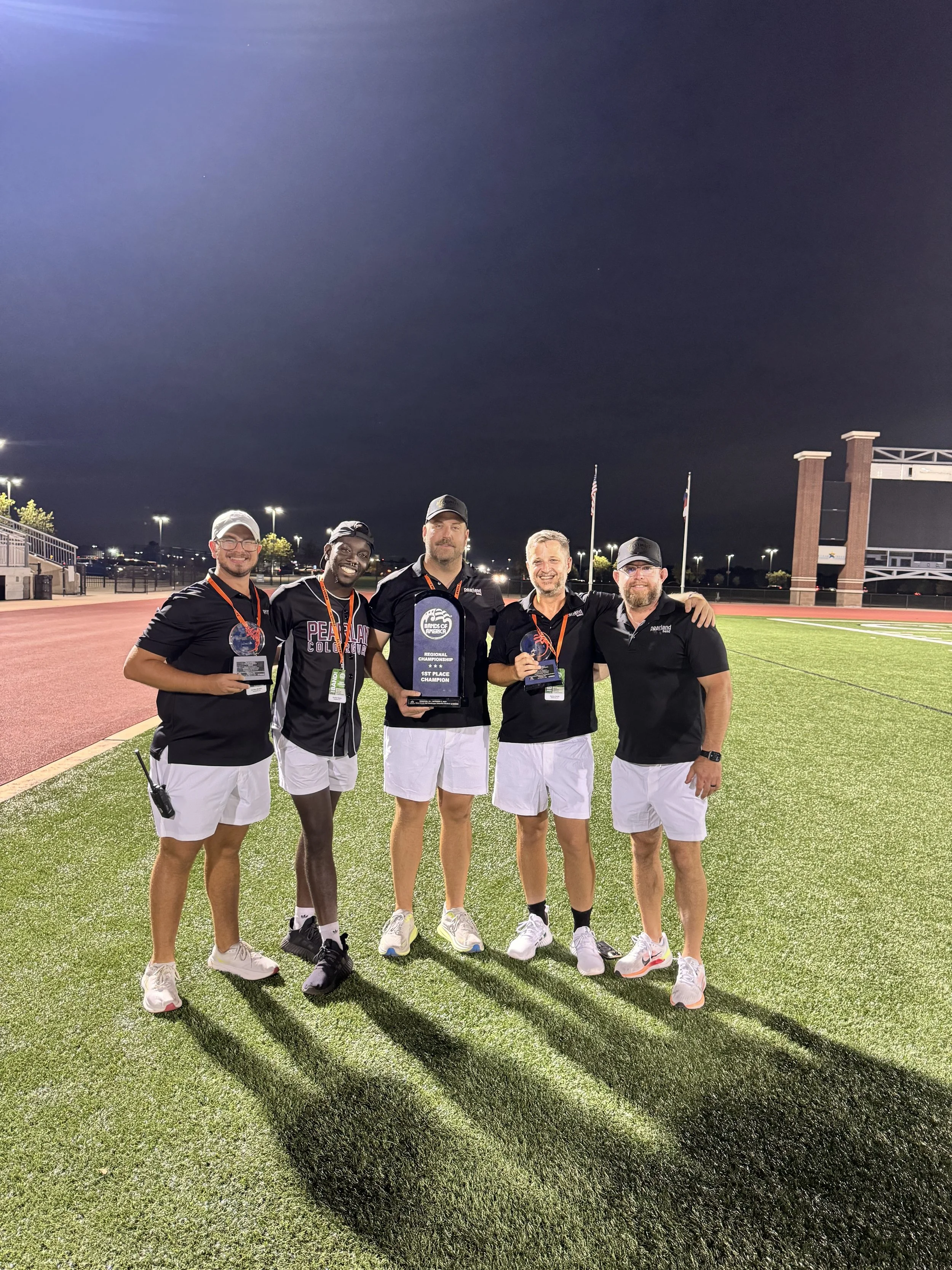 Group of five men holding trophies, standing on a sports field at night inside a stadium, celebrating a victory.