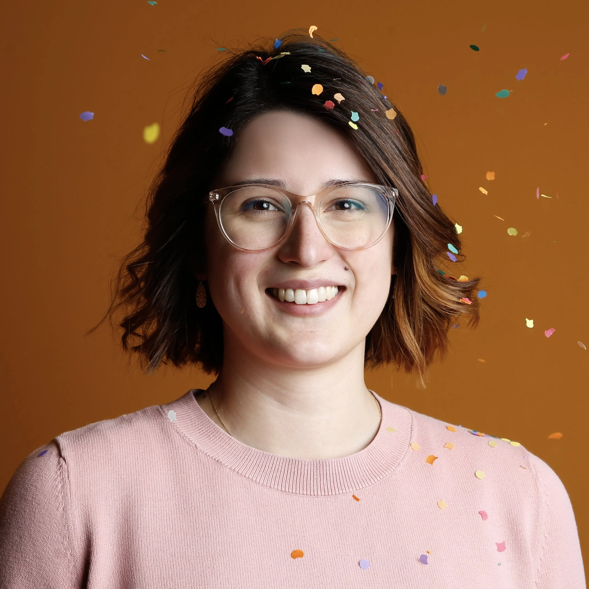 Eine lachende Frau trägt Brille, trägt ein pinkes Oberteil und ist von bunten Konfettistreifen umgeben, vor einem orangefarbenen Hintergrund.