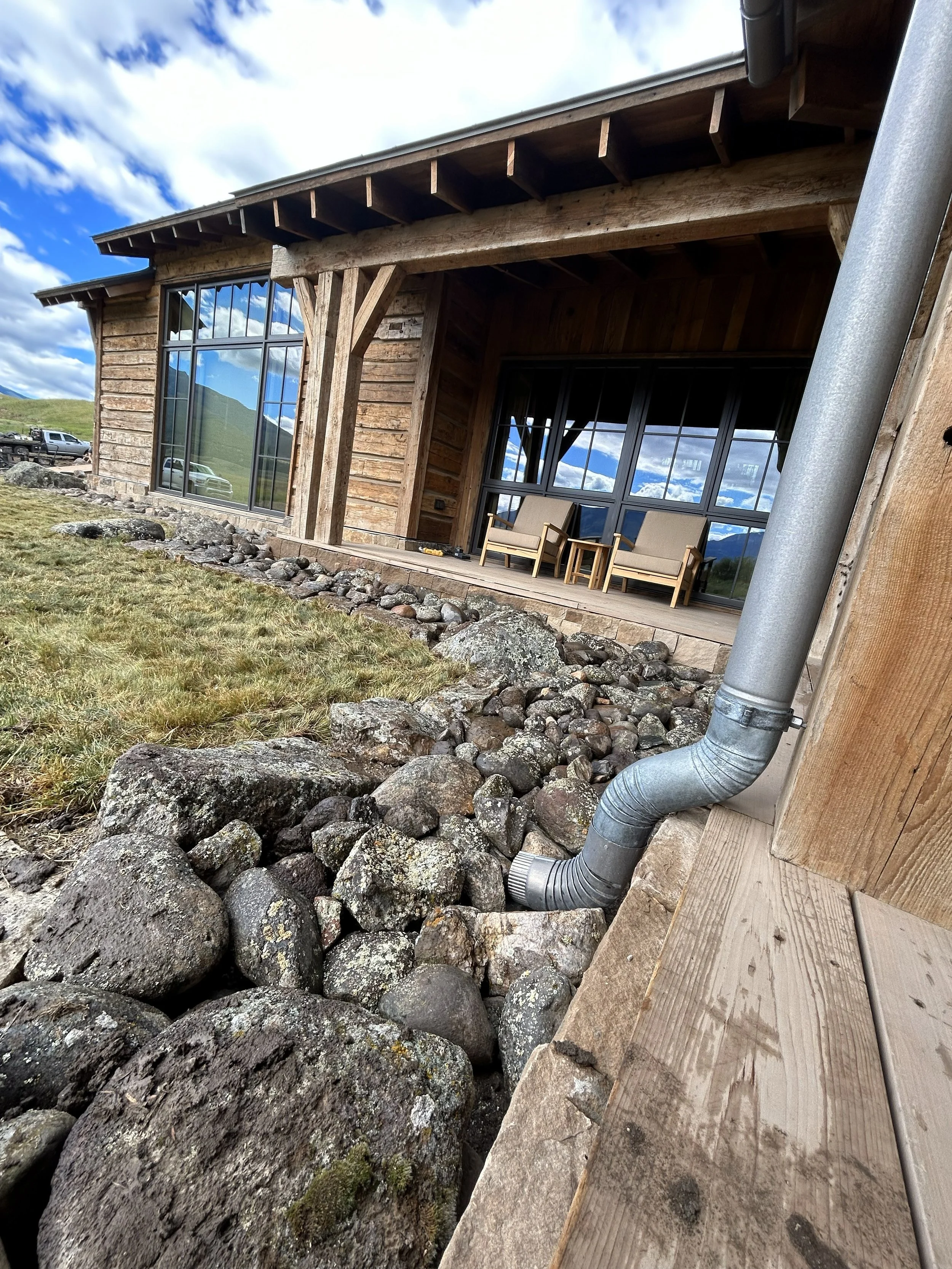 Close-up view of a building with large windows, wooden exterior, and a covered porch with outdoor furniture. Rocks and a metal downspout are in the foreground, with cloudy sky and mountains reflected in the windows.