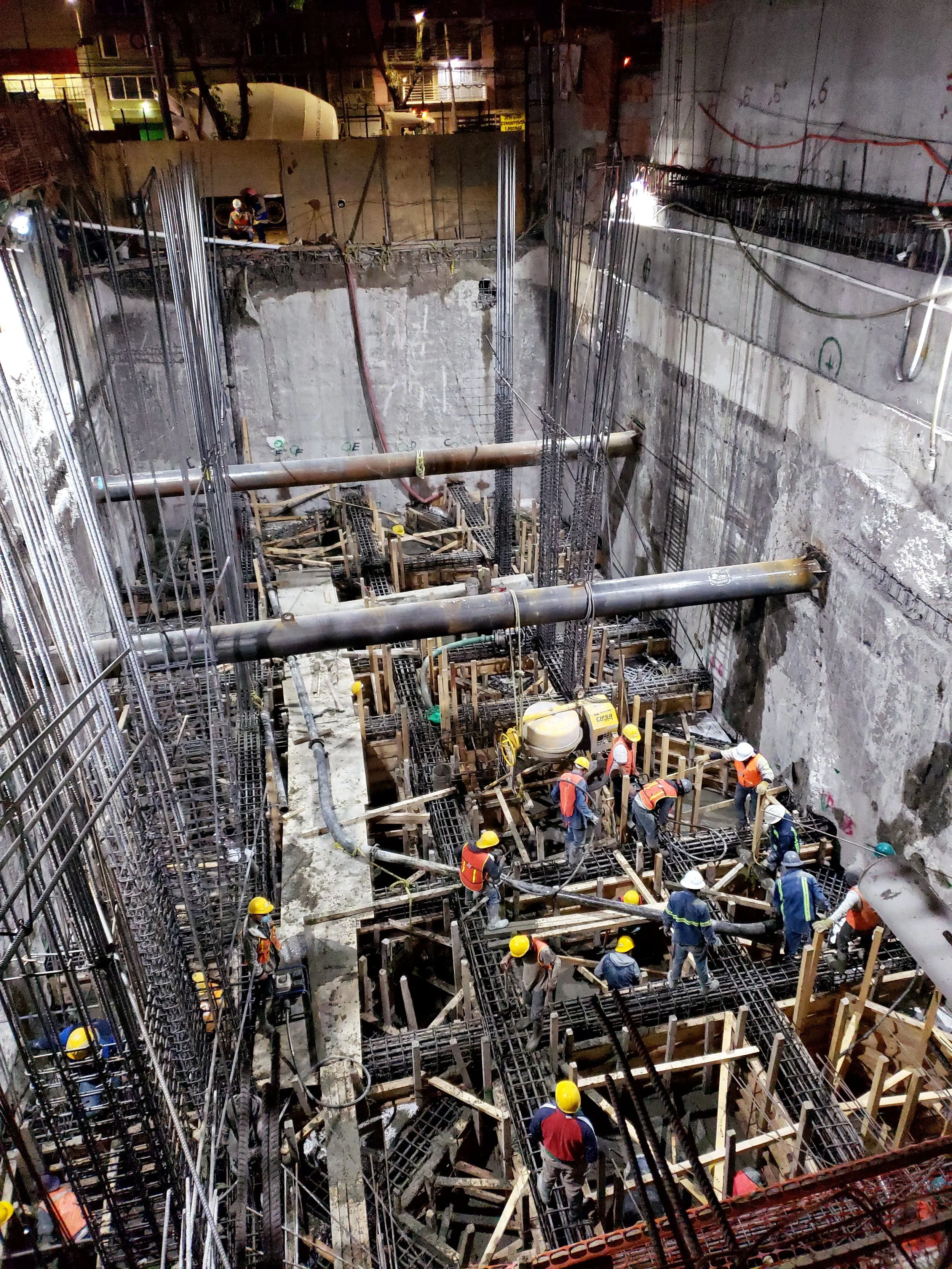 Construction workers in safety vests and helmets working on a deep construction site with steel rebar, scaffolding, and large pipes.