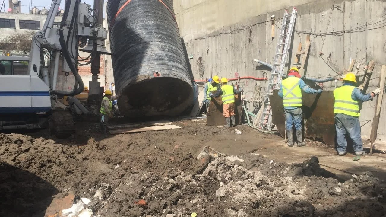 Construction workers with yellow safety vests and helmets working on a construction site. Large black pipe being lowered into place beside a concrete wall. Construction equipment, ladder, and dirt are visible.