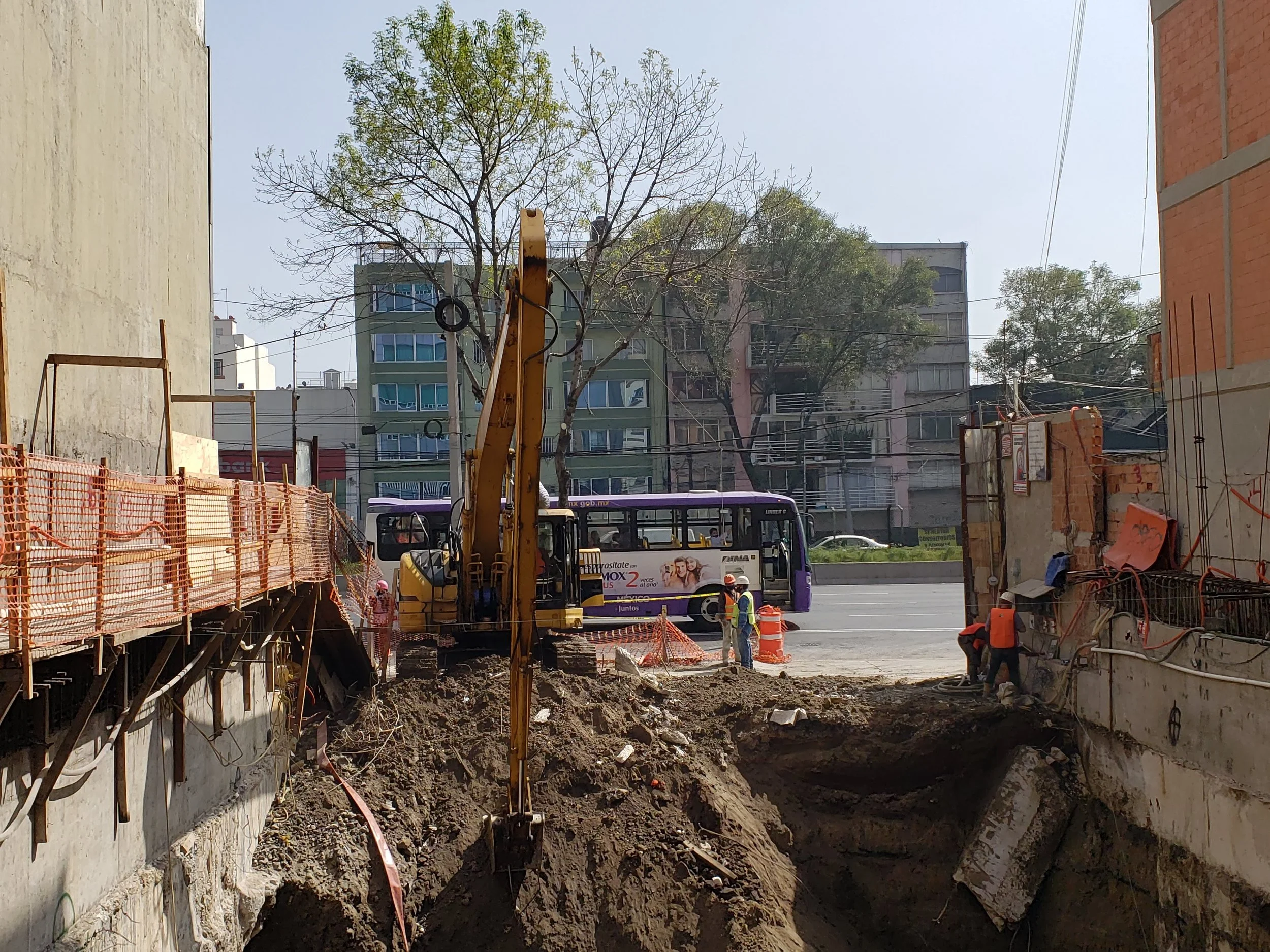 Construction site with workers and excavator near a street, with a bus and residential buildings in the background.