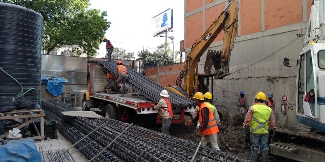 Construction workers on a site with steel rebar, a crane, and a dump truck with workers unloading materials.