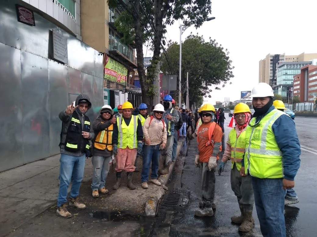 Street workers and construction crew standing on the sidewalk during a road repair project, wearing safety gear such as helmets and vests, with buildings and cars in the background.