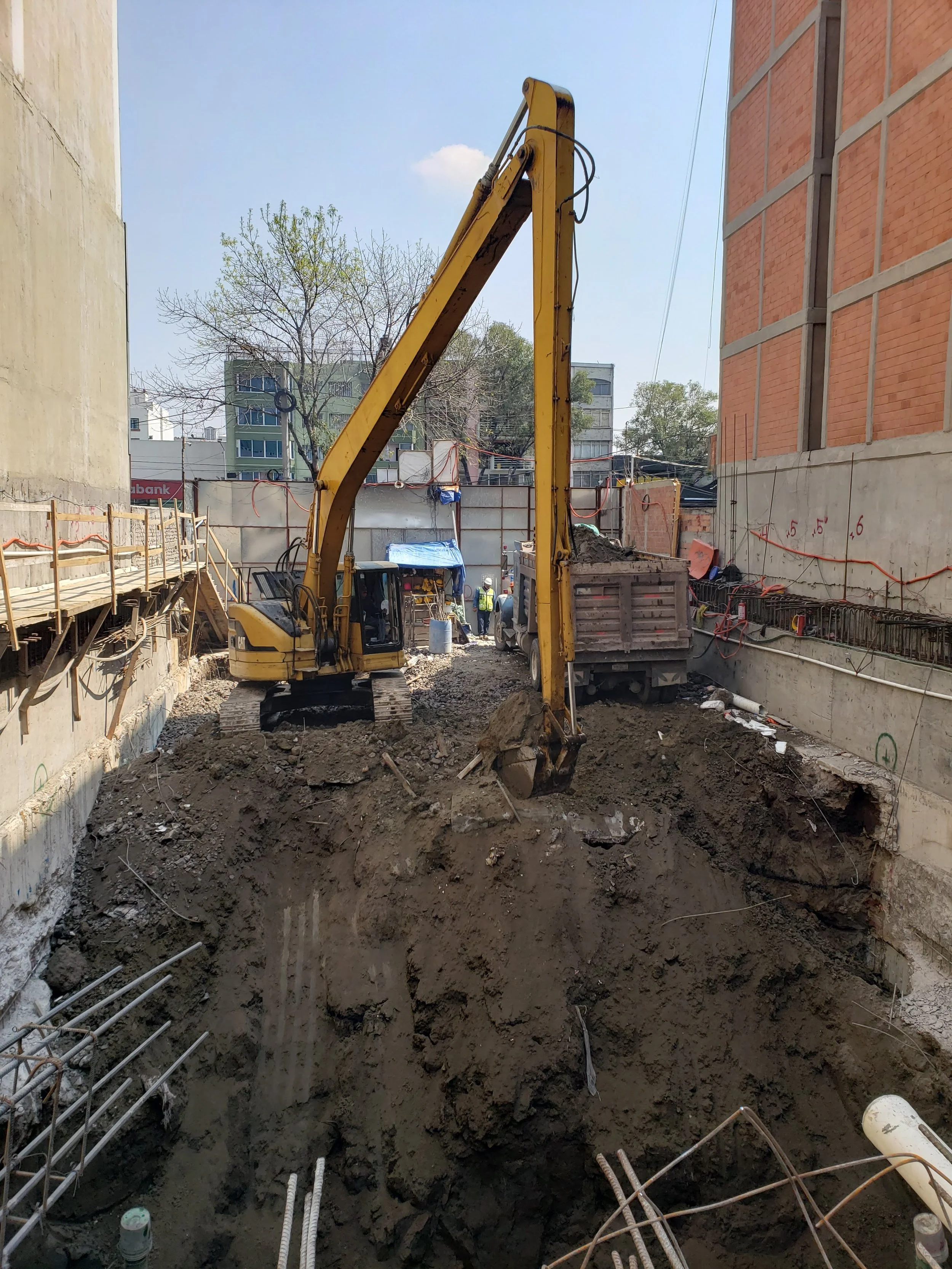 Construction site with excavator digging a trench, construction workers in the background, surrounded by building walls and construction materials.