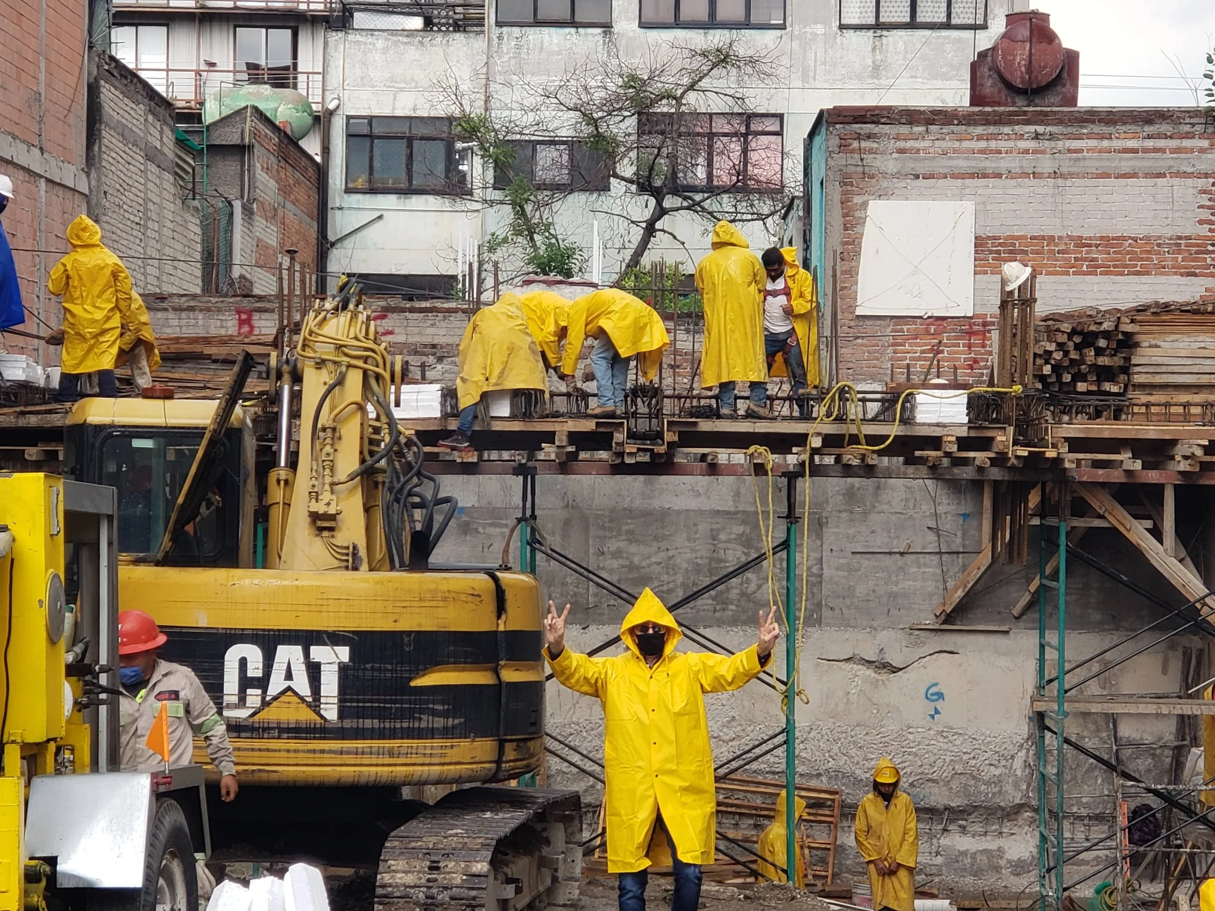 Construction workers in yellow raincoats working on a building site, with a person in a yellow raincoat and sunglasses giving a peace sign in front of a yellow CAT excavator.
