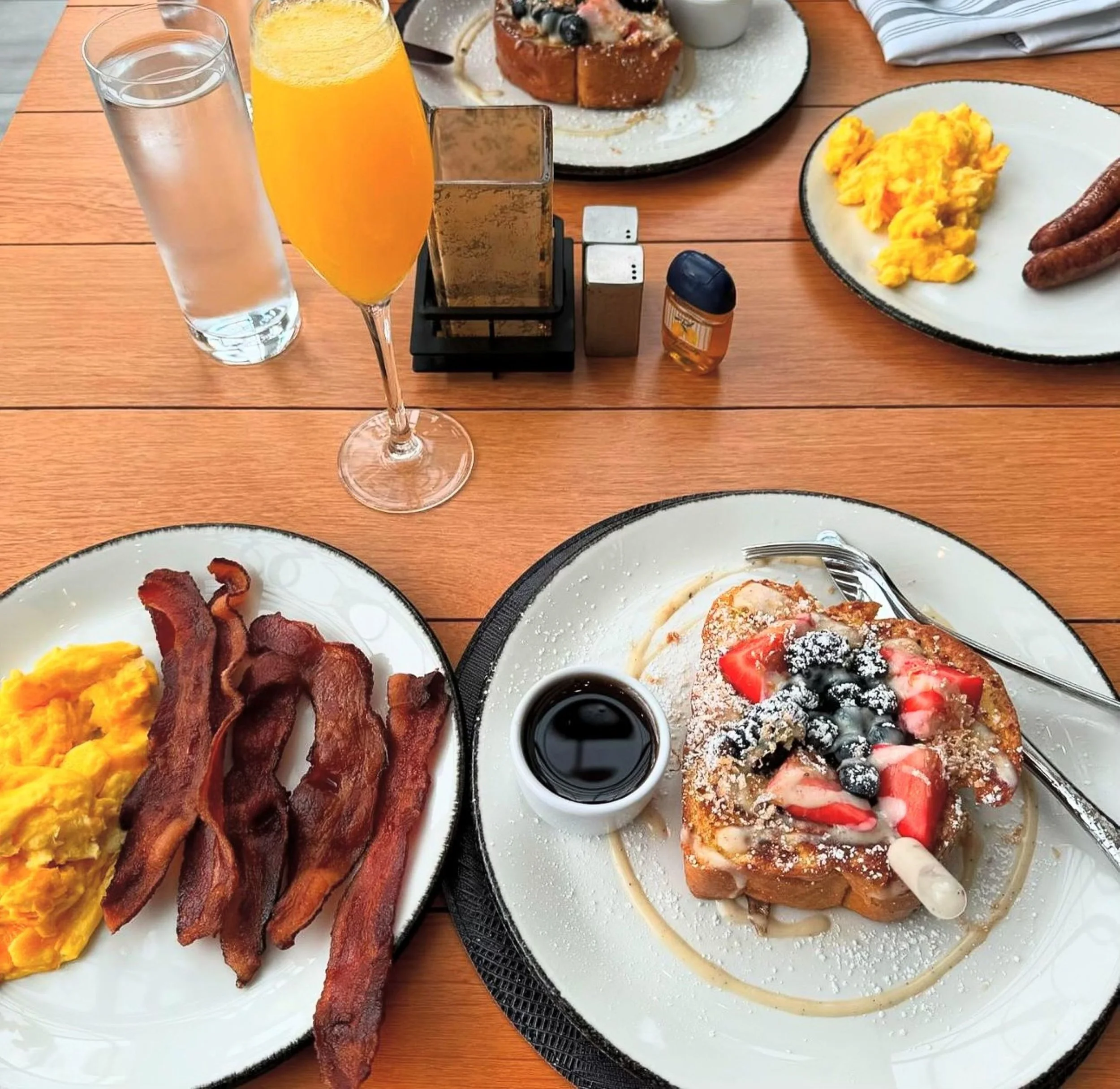 Breakfast with scrambled eggs, bacon, French toast with fruit topping and syrup, a glass of orange juice, a glass of water, and a glass of soda on a wooden table.