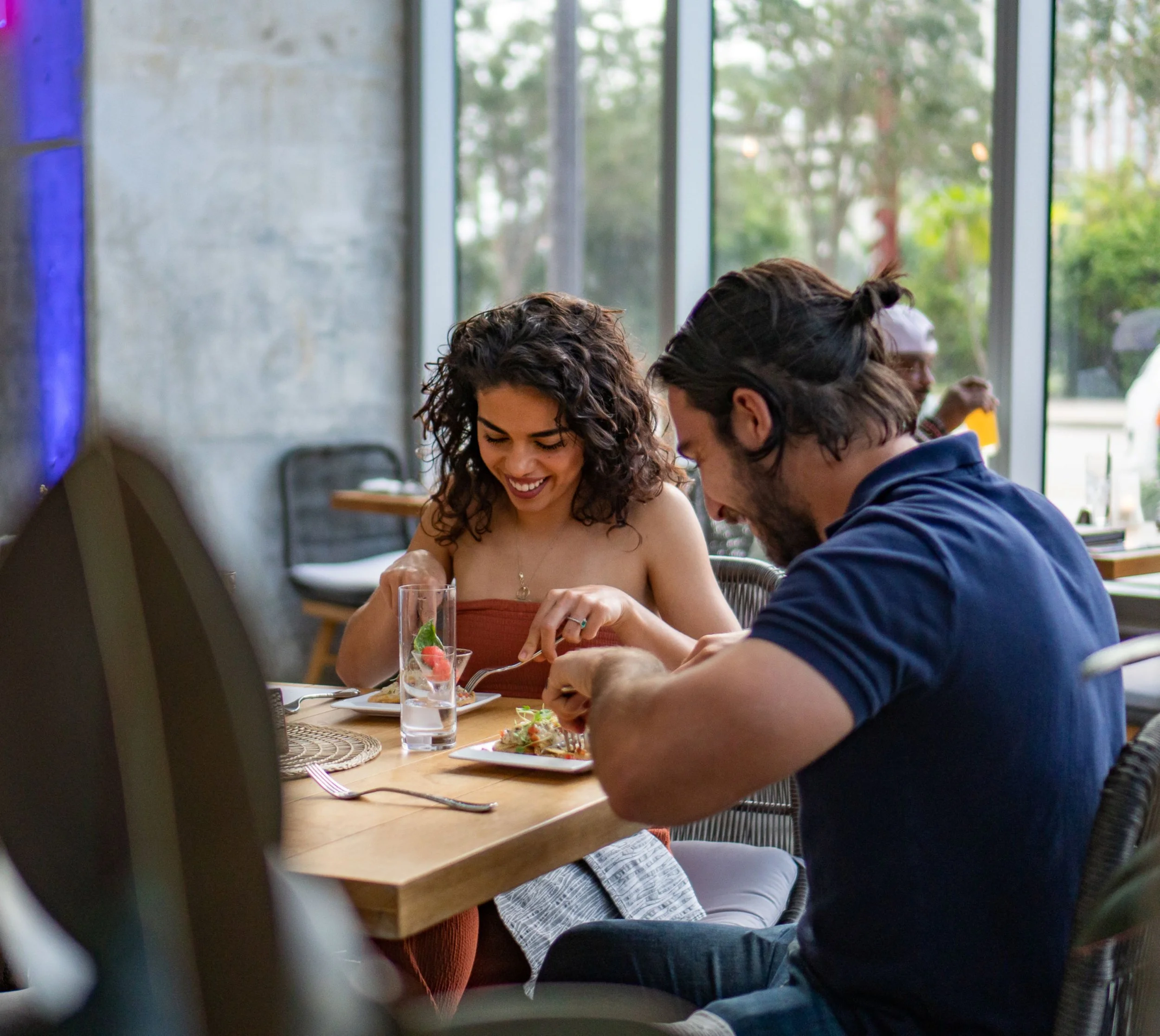 A smiling woman and man enjoying a meal at a restaurant, with large windows and city greenery in the background.