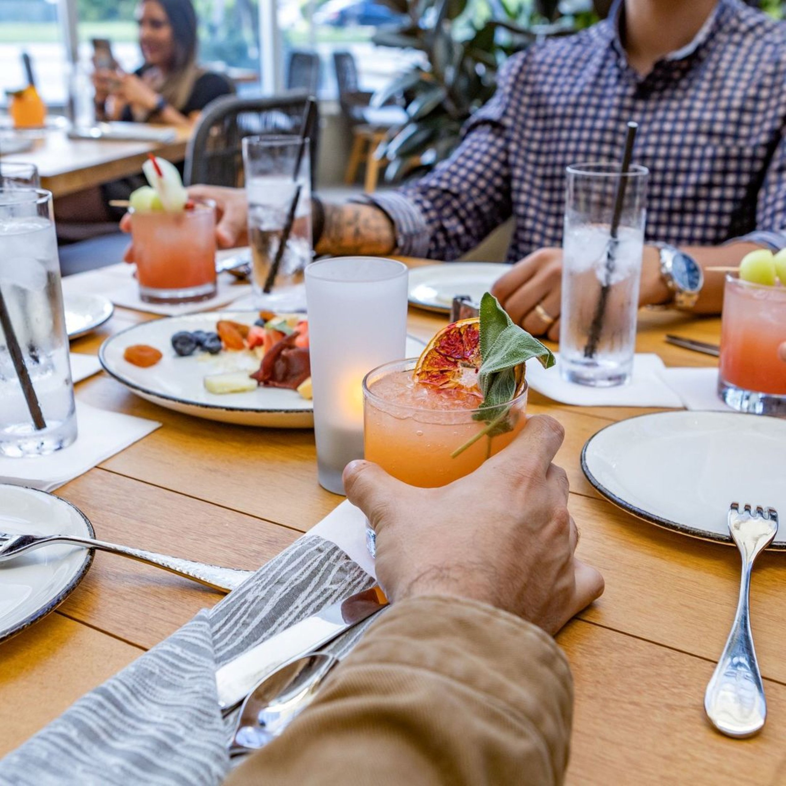 A group of people gathered around a dining table enjoying drinks and food at a restaurant or cafe. A hand in the foreground holds a cocktail garnished with a slice of dried orange and a leaf.