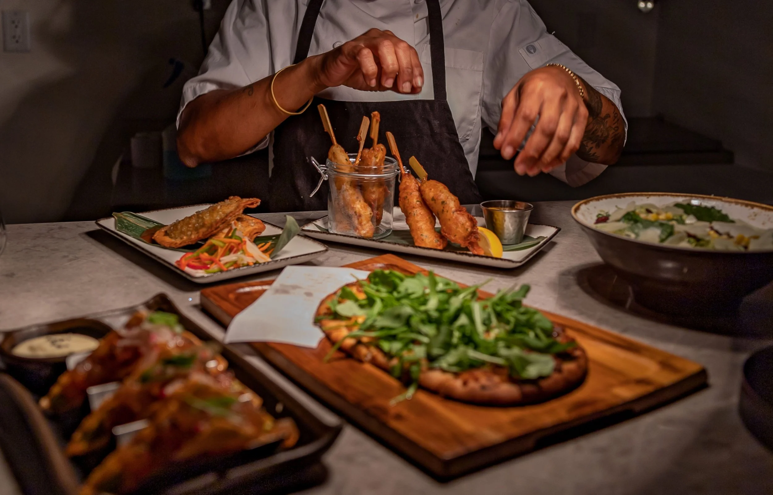 Chef garnishing plates of fried appetizers, like skewers, in a dimly lit restaurant with various dishes on the table, including pizza, salad, and a bowl of greens.