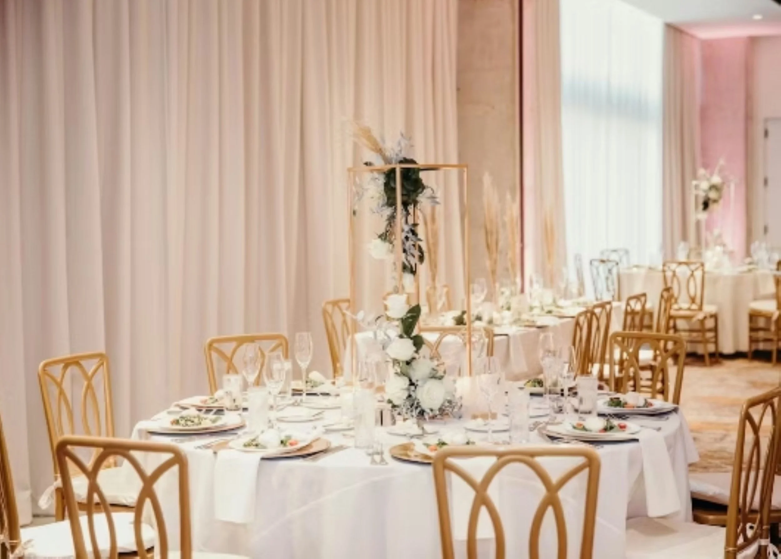 Elegant banquet table set for a celebration or wedding with white tablecloth, floral centerpiece, glasses, plates, and chairs in a decorated room with draped curtains and soft lighting.