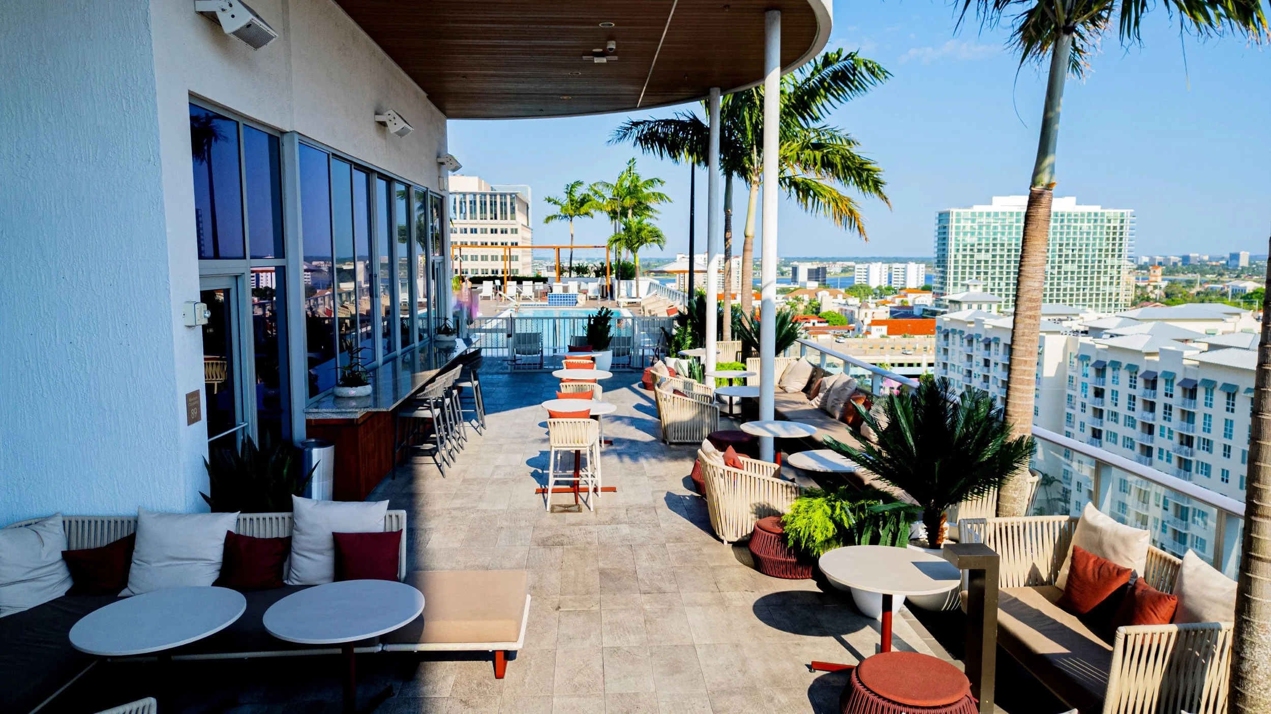 Outdoor rooftop patio with lounge chairs, tables, and palm trees, offering a city view with buildings and blue sky.