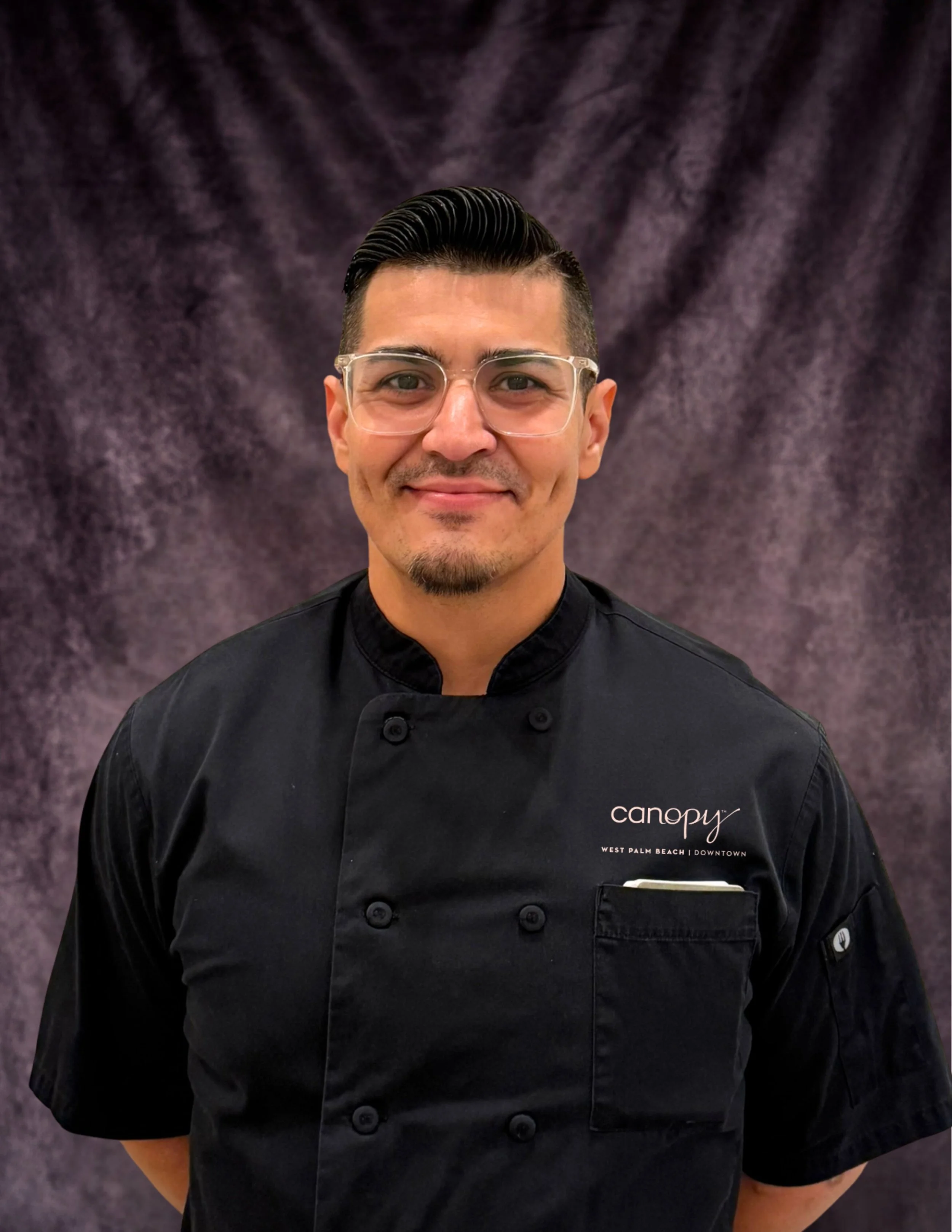 A man with glasses and styled dark hair wearing a black chef coat with 'Canopy' logo and text 'West Palm Beach | Downtown' standing in front of a textured gray background.