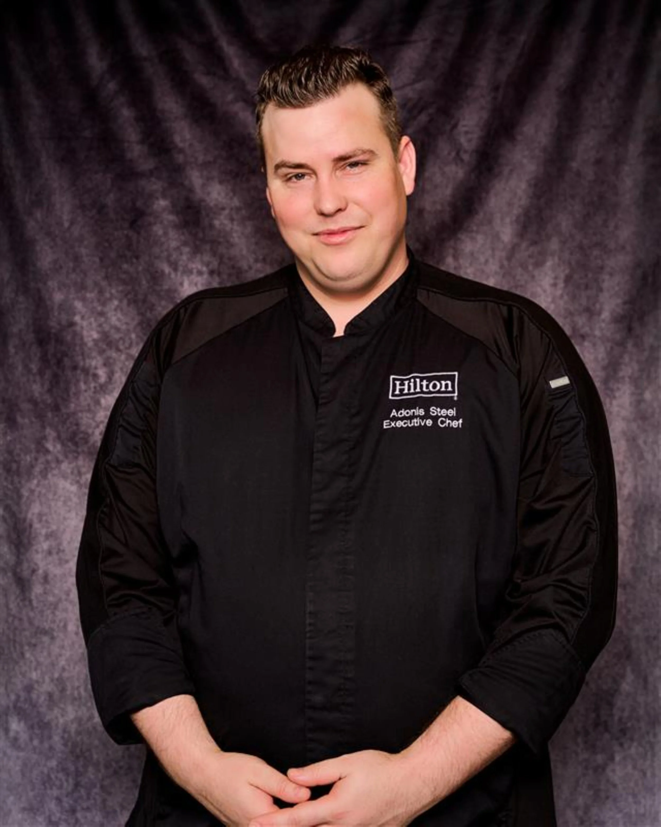 A man in a black chef's coat with Hilton logo and text that reads 'Adonis Steel, Executive Chef' standing against a gray textured background.