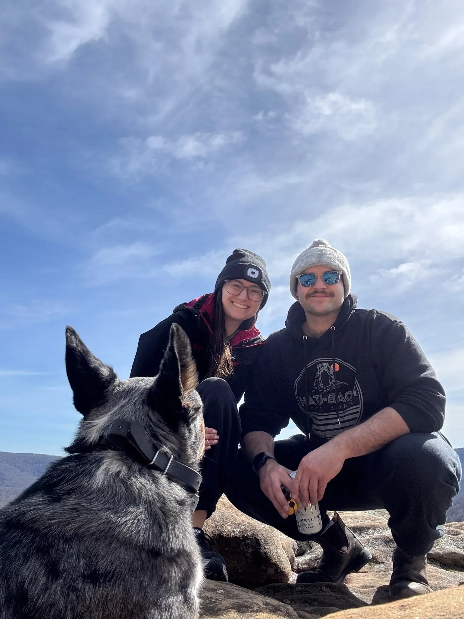 A smiling woman and man in outdoor clothing with a dog on a rocky landscape under a partly cloudy sky.
