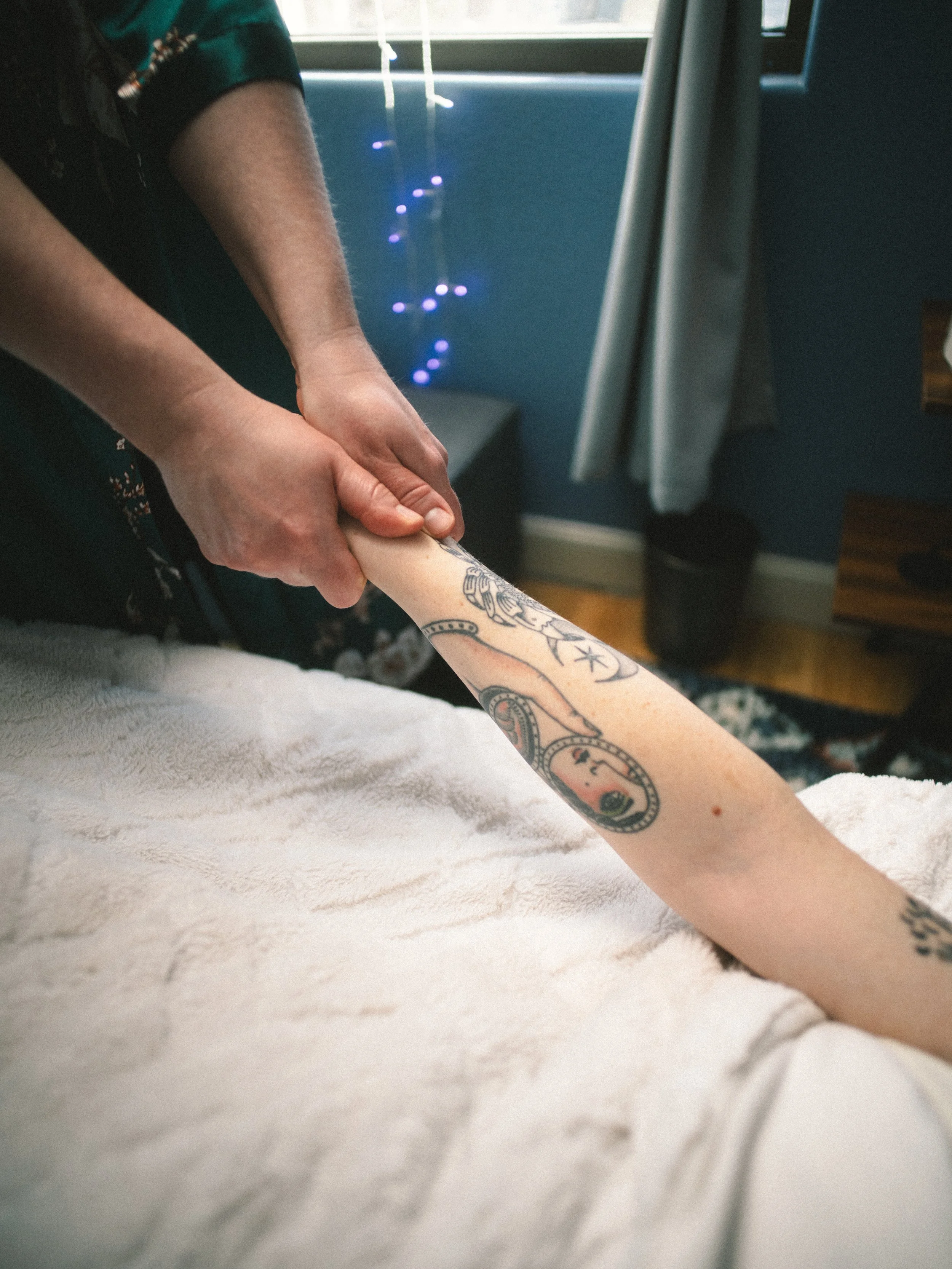 A person receiving a massage or physical therapy on their arm, with a focus on the tattooed forearm against a bedroom background with a window and blue wall.
