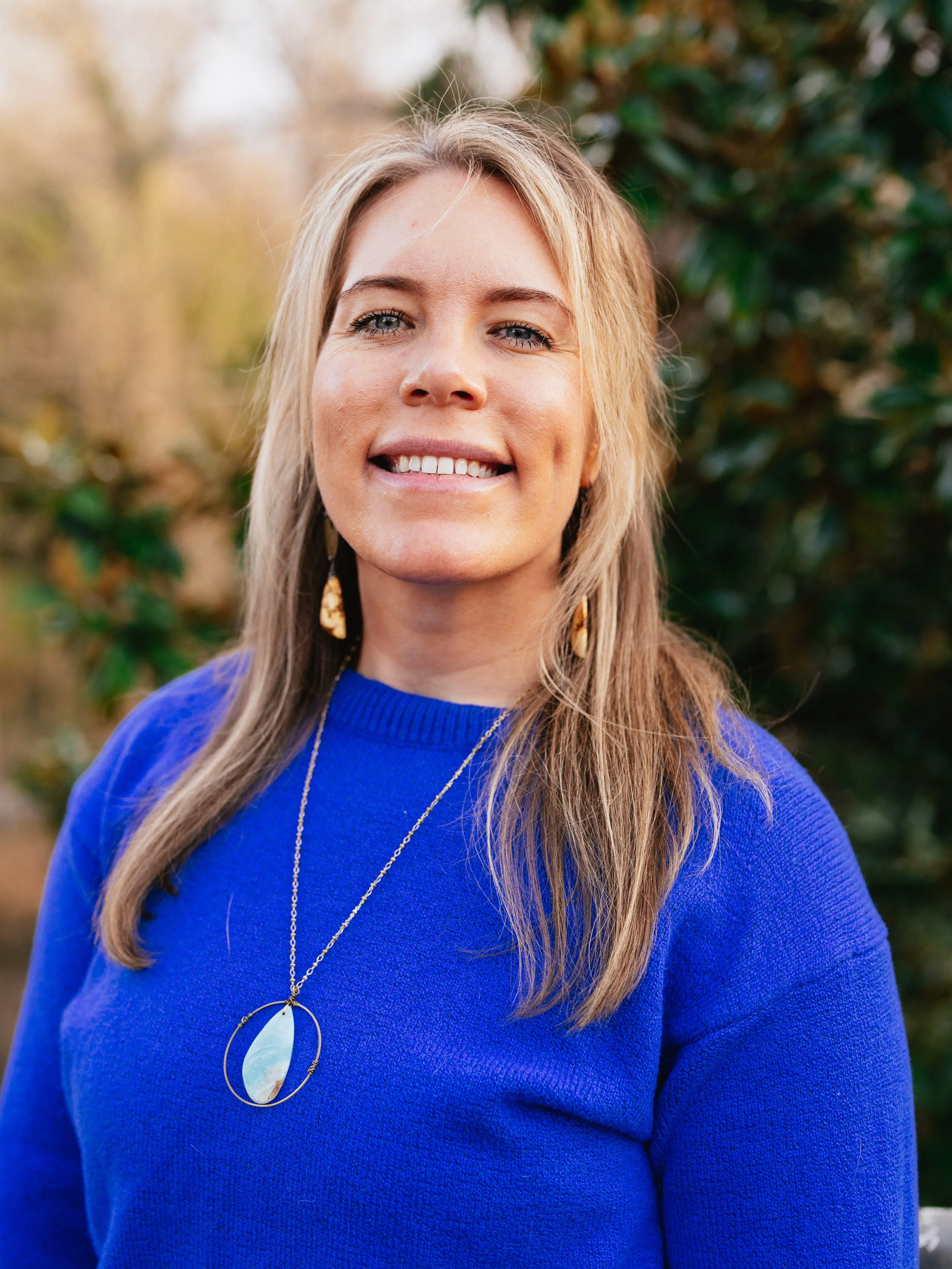 A woman smiling outdoors, wearing a bright blue sweater and a necklace, with green foliage in the background.