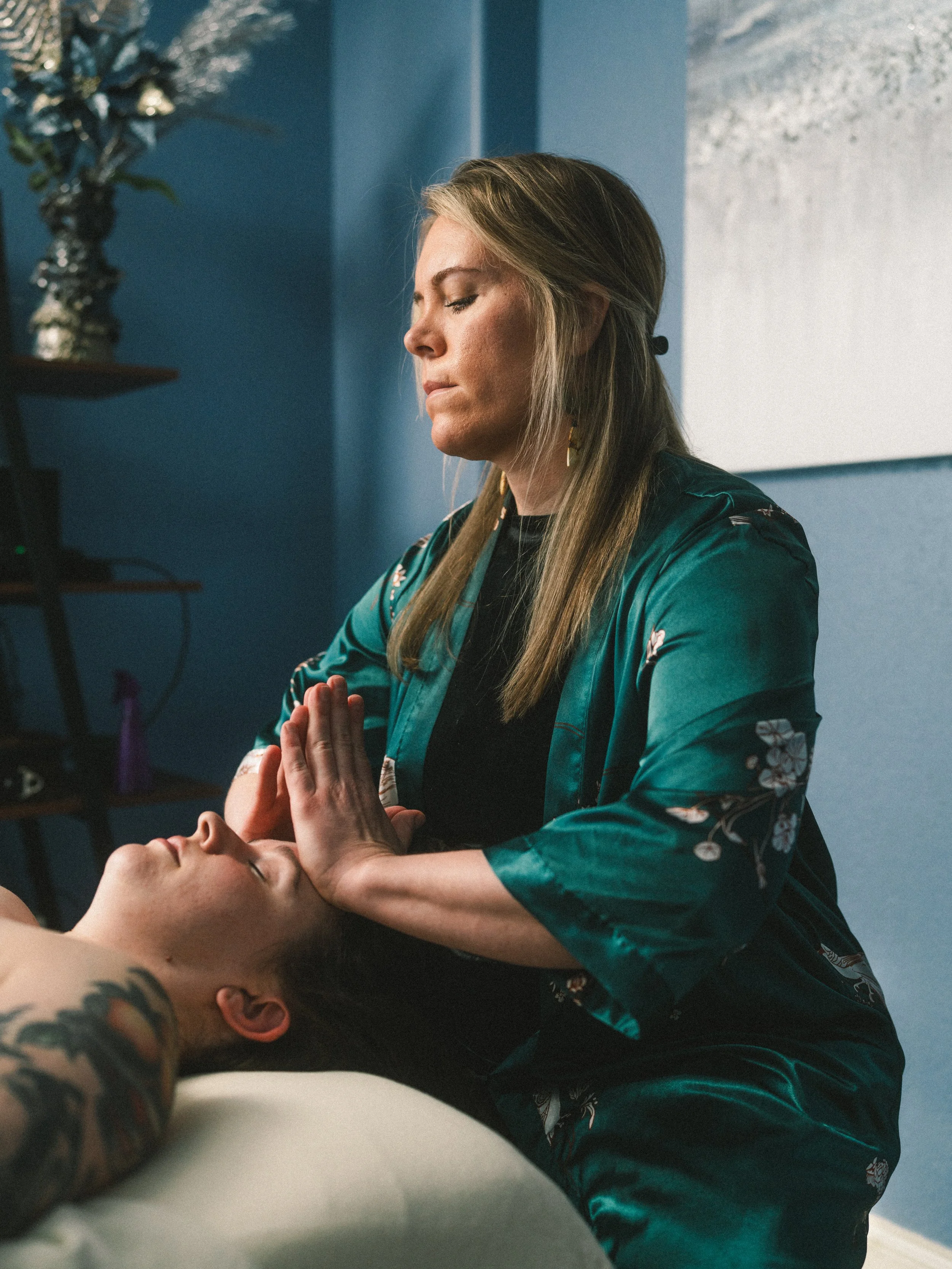 A woman performs a healing or massage technique on a man lying on a bed, with her hands pressed together in a prayer-like position near his forehead.
