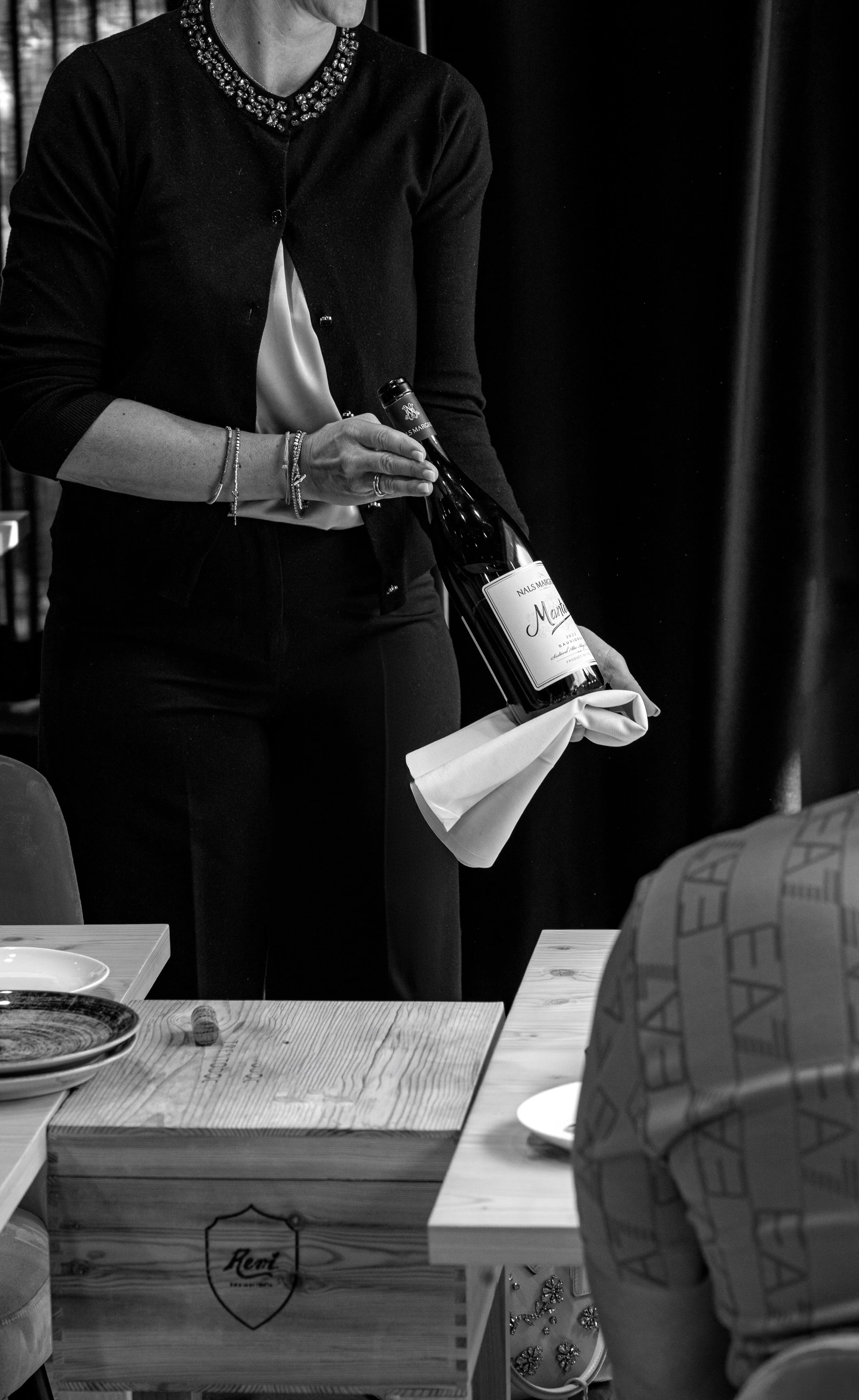 Una donna sta consegnando una bottiglia di vino durante un evento. Si vedono anche altre persone e piatti sul tavolo.