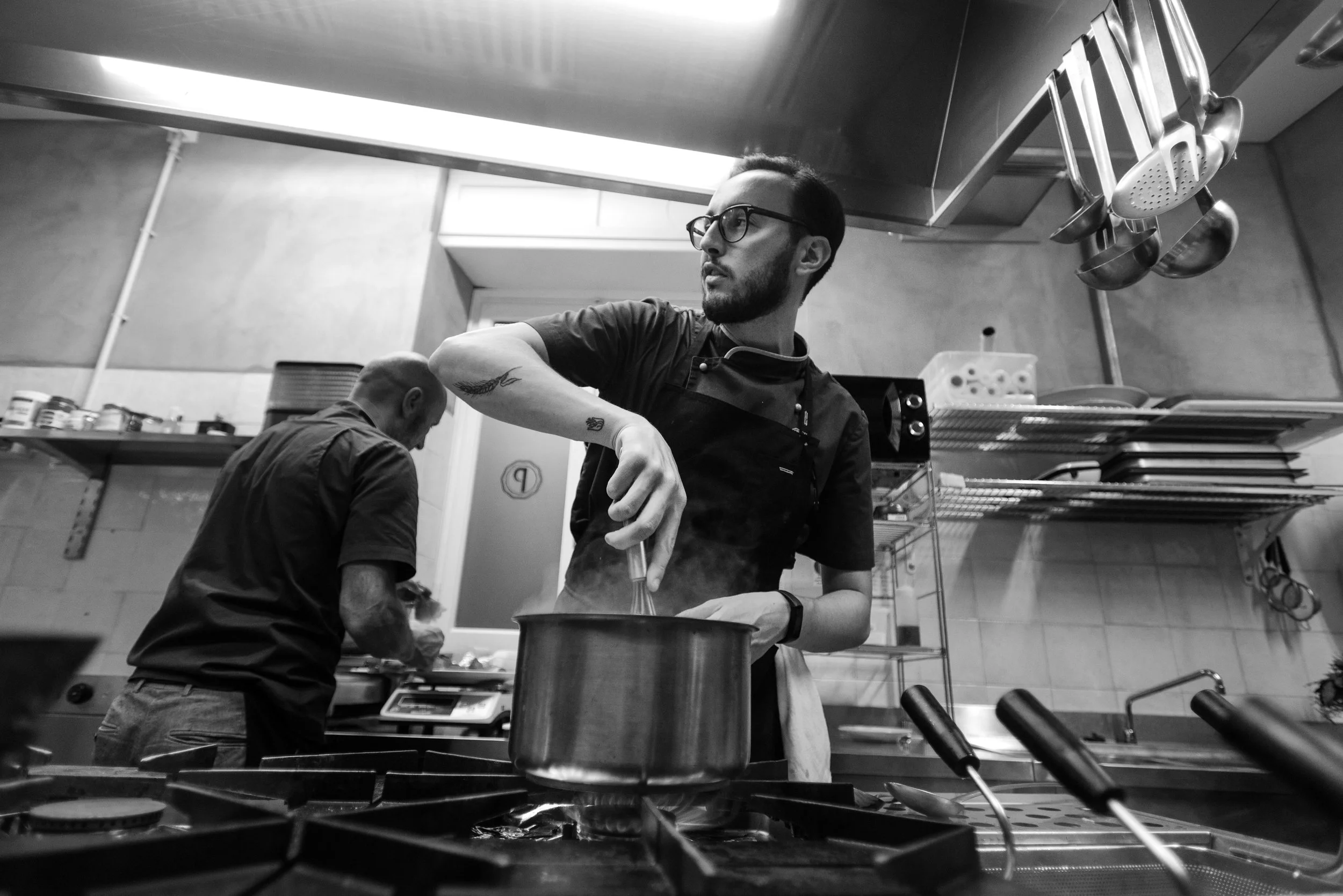 Uomo con occhiali e barba che cucina in cucina professionale, attivo su fornelli, con altri chef sullo sfondo in un ambiente di cucina industriale.