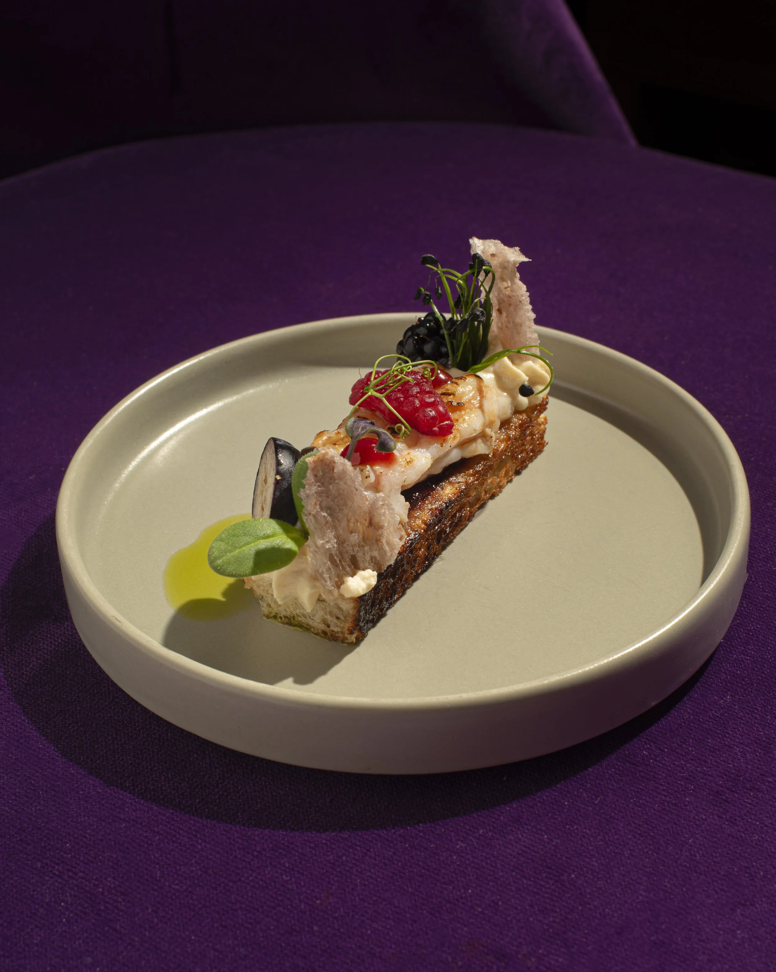 Piatti gourmet con diversi ingredienti, decorati con erbe e accompagnati da una salsa verde, presentati su una pirofila bianca su sfondo viola.