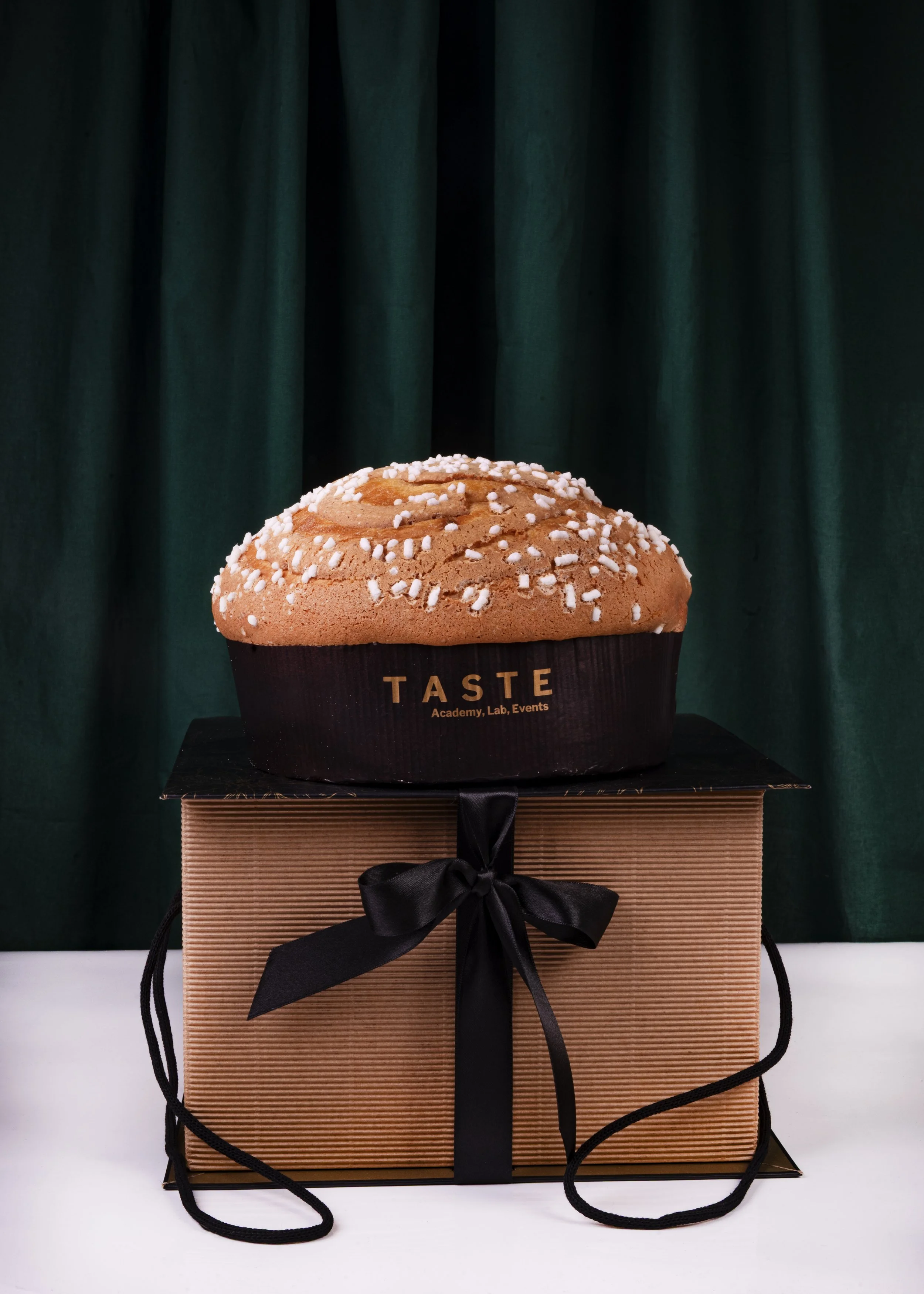 Una grande torta con crema color caramello e zuccherini bianchi decorativi, posizionata su una scatola di cartone marrone con fiocco nero e confezione nera con scritto 'Taste' e 'Academy, Lab, Events'