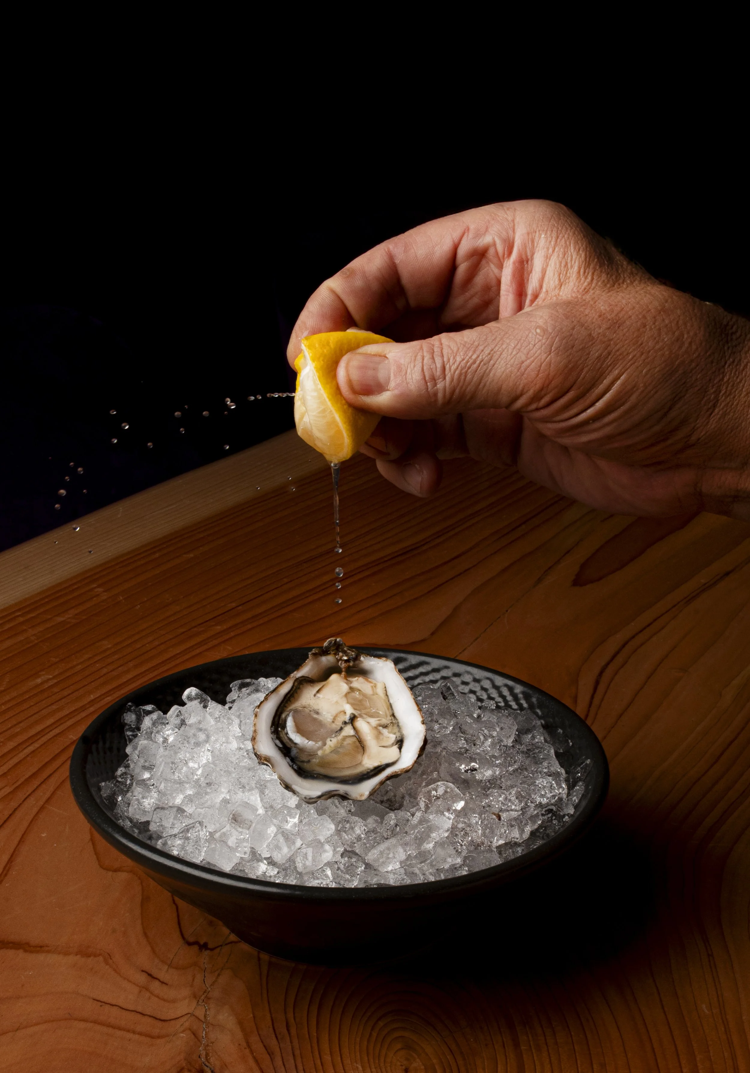 Una mano stringe un limone tagliato, con alcune gocce di succo che cadono su un piatto di ostriche su ghiaccio, con uno sfondo nero e un piano di legno.