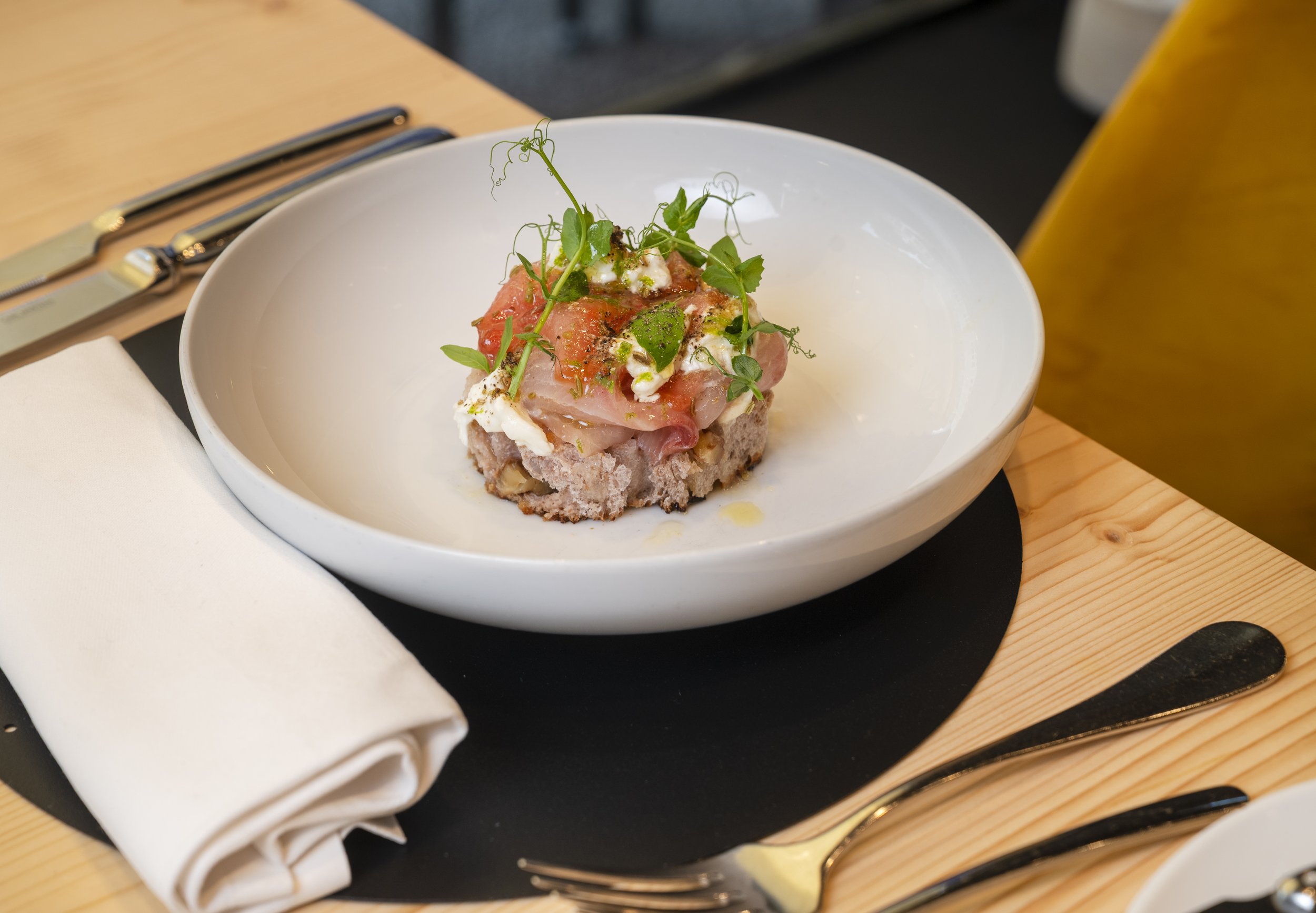 Piattino con tartare di carne decorata con germogli e erbe aromatiche, accompagnato da coltelli, forchetta e tovaglioli bianco su un tavolo di legno.