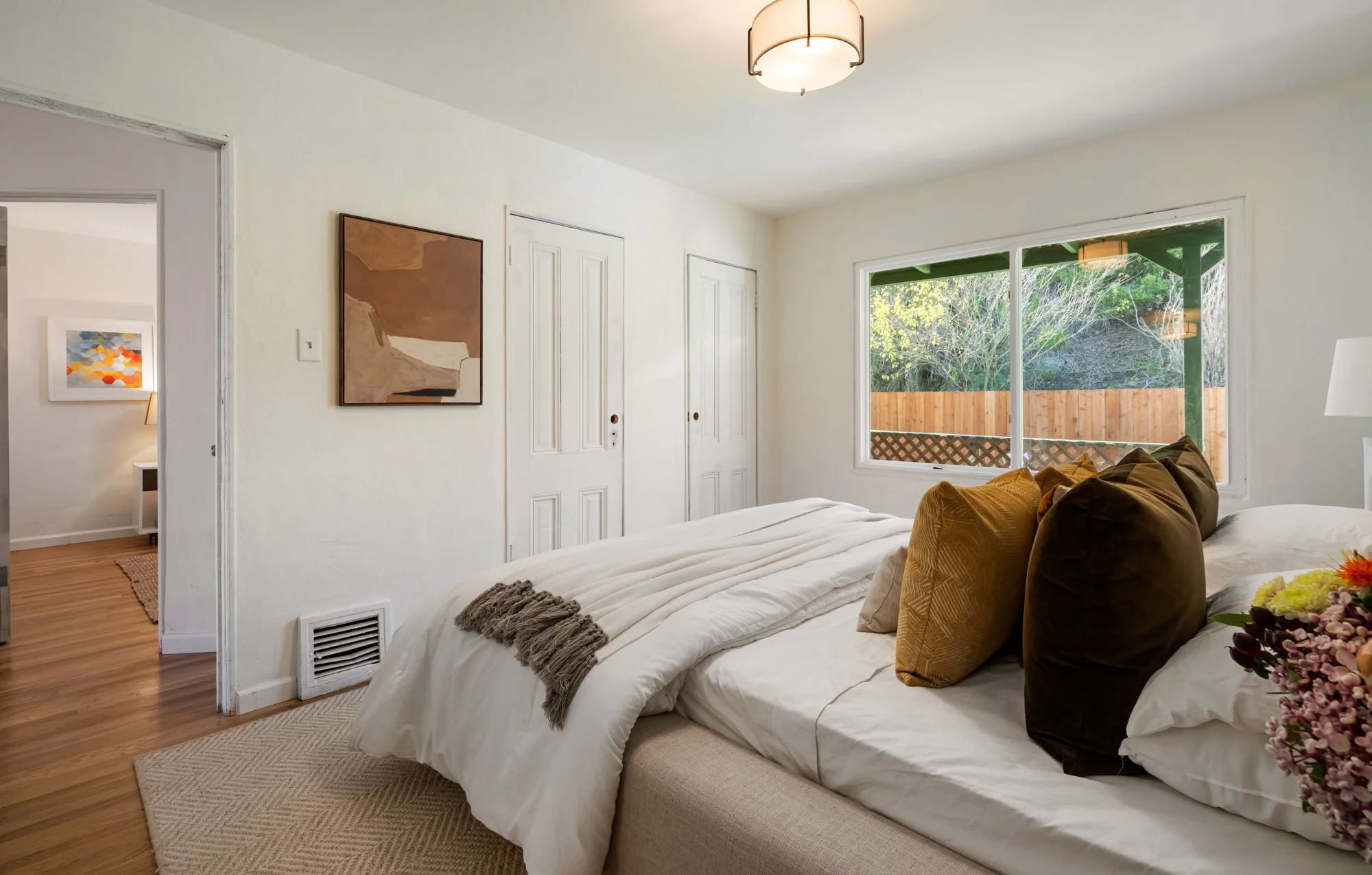 A bedroom with a large bed, multiple pillows, a window showing a wooden fence and greenery outside, and a closet with double doors.