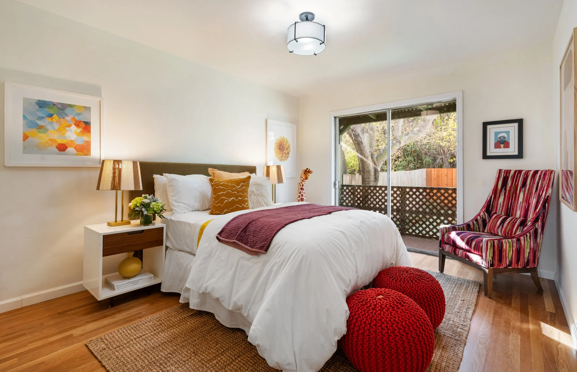 A cozy bedroom with a white bed, colorful pillows, two lamps, and a sliding glass door leading to a small balcony with trees visible outside.