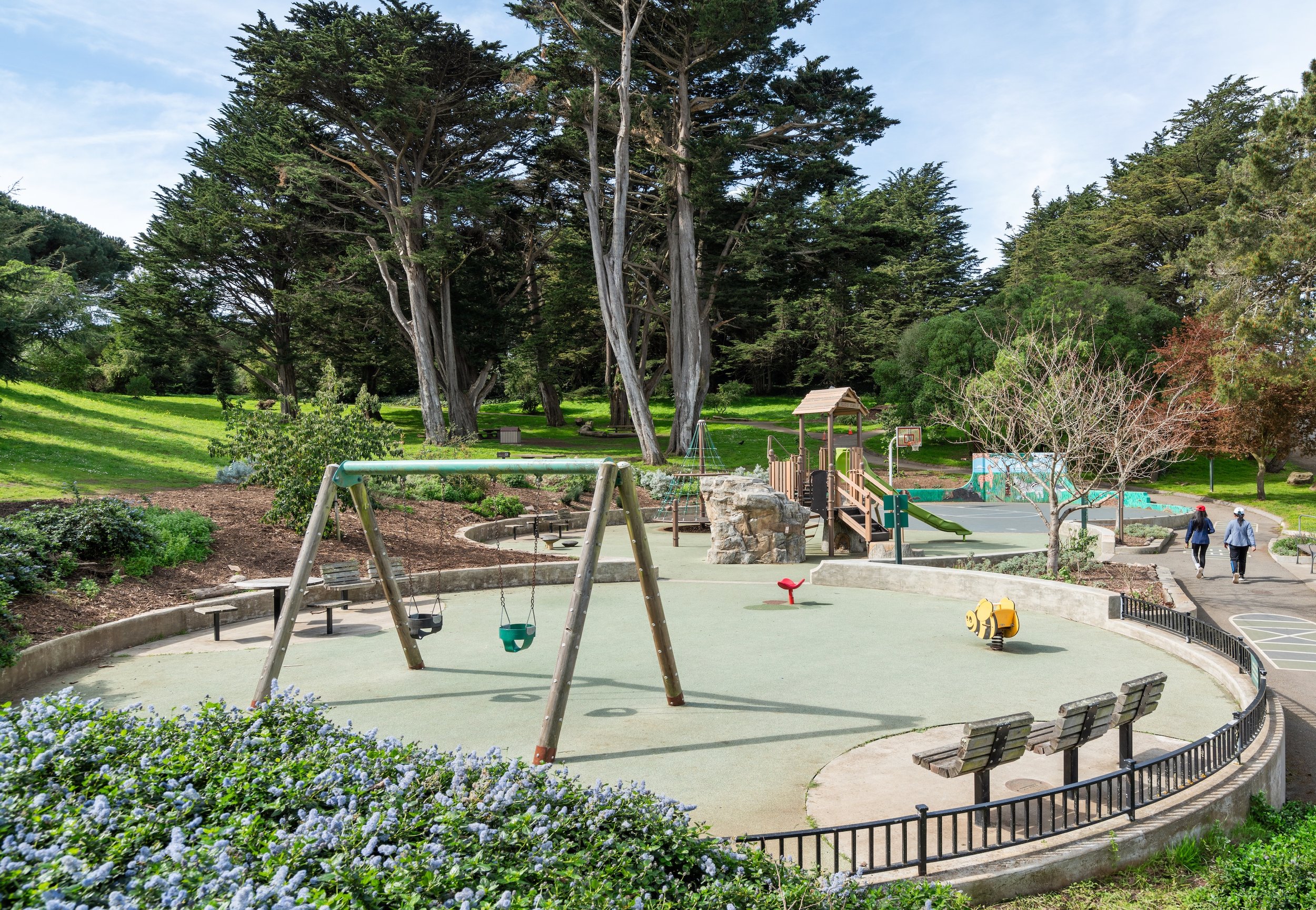 A playground in a park with swings, spring rider, slide, and basketball hoop, surrounded by trees and benches, with two people walking in the background.