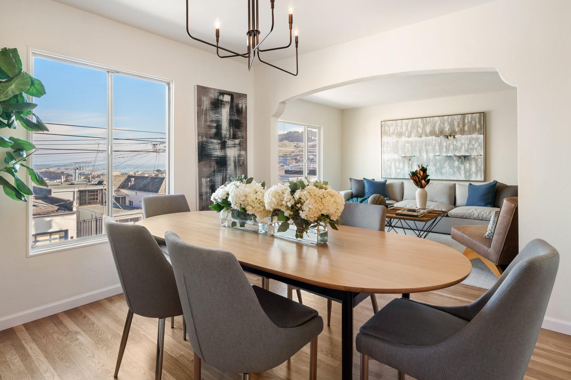 Dining room with wooden table, six gray chairs, floral centerpiece, and large windows showing city views; living room with sofa, armchair, large abstract artwork, and decorative pillows.