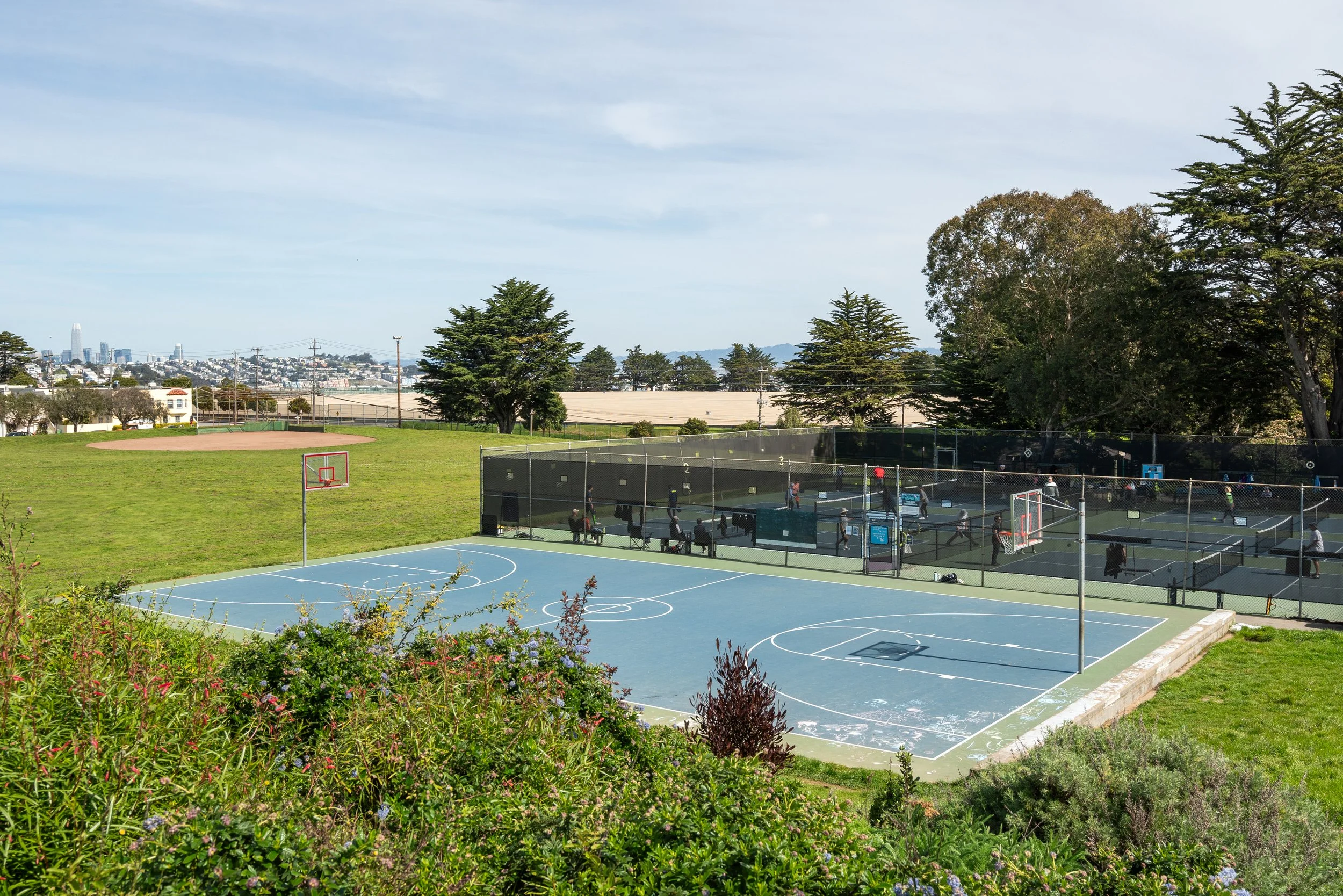 A recreational area with a basketball court and several tennis courts enclosed by a fence, with people playing tennis and sitting nearby. There is greenery with trees and shrubs, and a city skyline in the background under a partly cloudy sky.