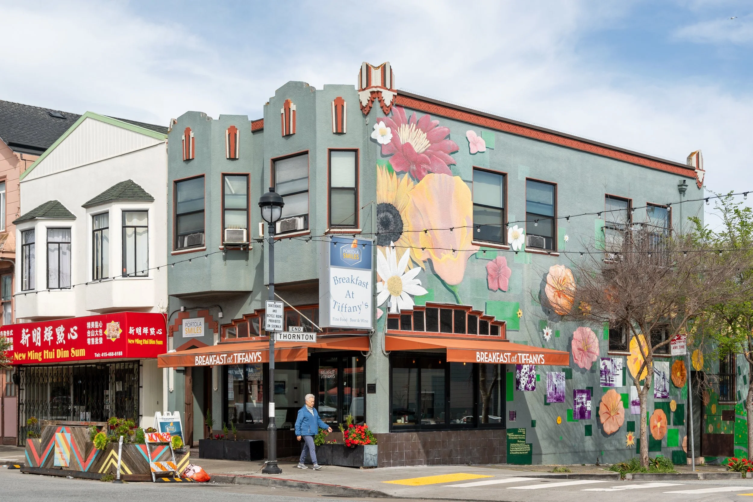 A three-story building with colorful murals of flowers on its exterior wall, featuring businesses including a breakfast cafe named Tiffany's and a dim sum restaurant with red signage. There's a woman walking on the sidewalk and some trees and street 