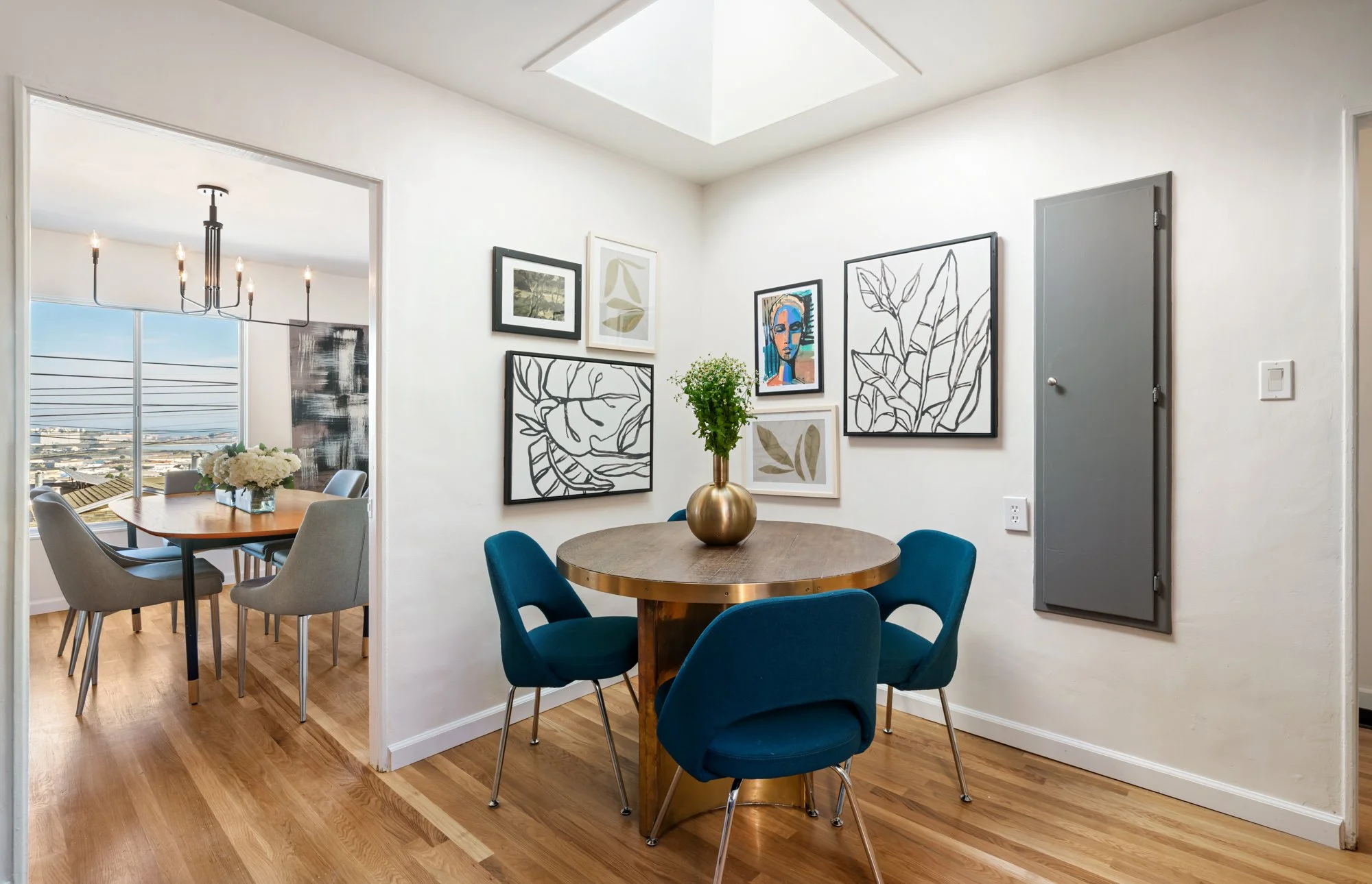 Dining area with a round wooden table, teal chairs, a gold vase with green plants, and wall art, with a view of the adjacent room with a chandelier and large window.