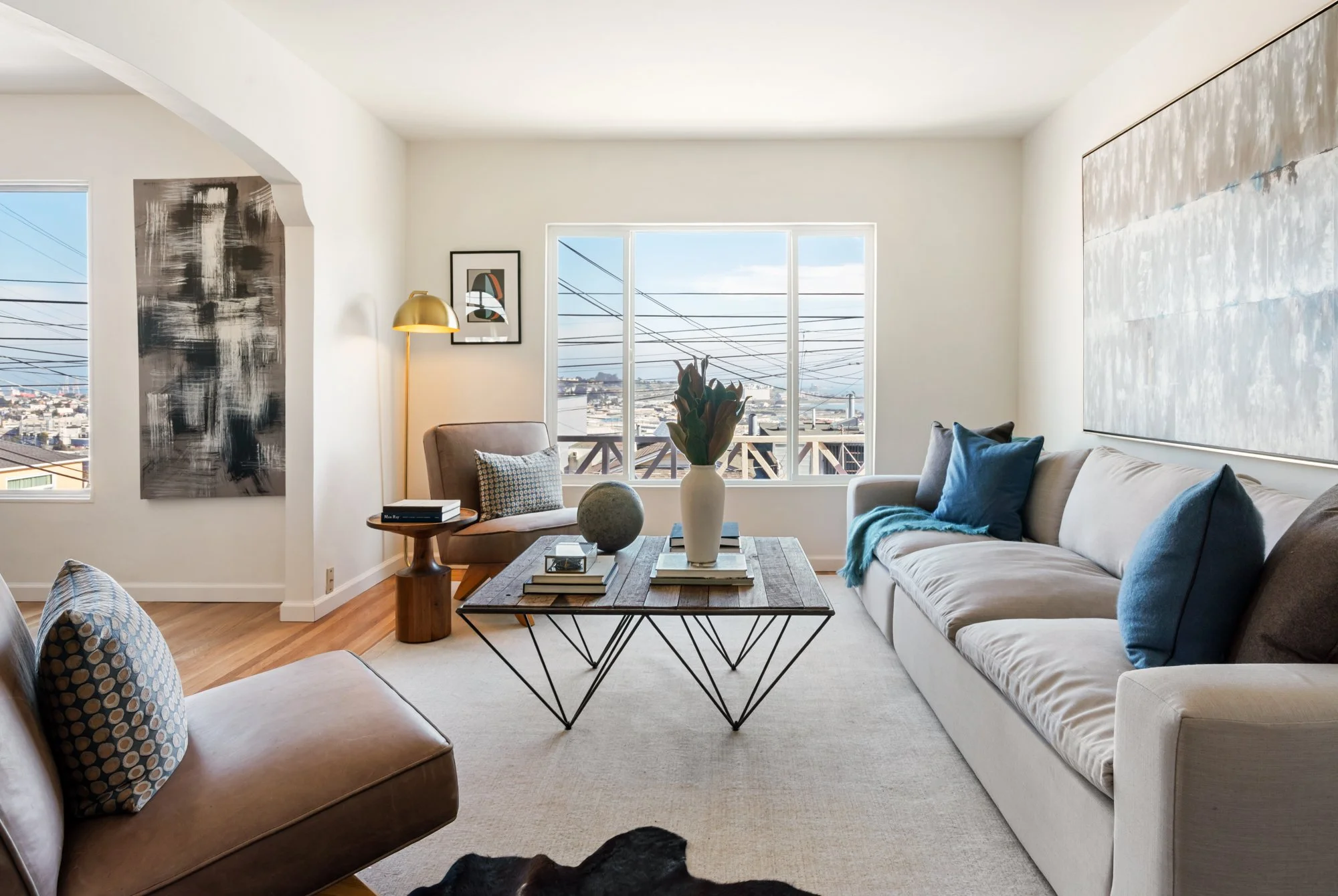 Living room with beige sofa, blue pillows, a coffee table with a vase and books, a tan armchair, abstract wall art, and large windows showing a cityscape with power lines.