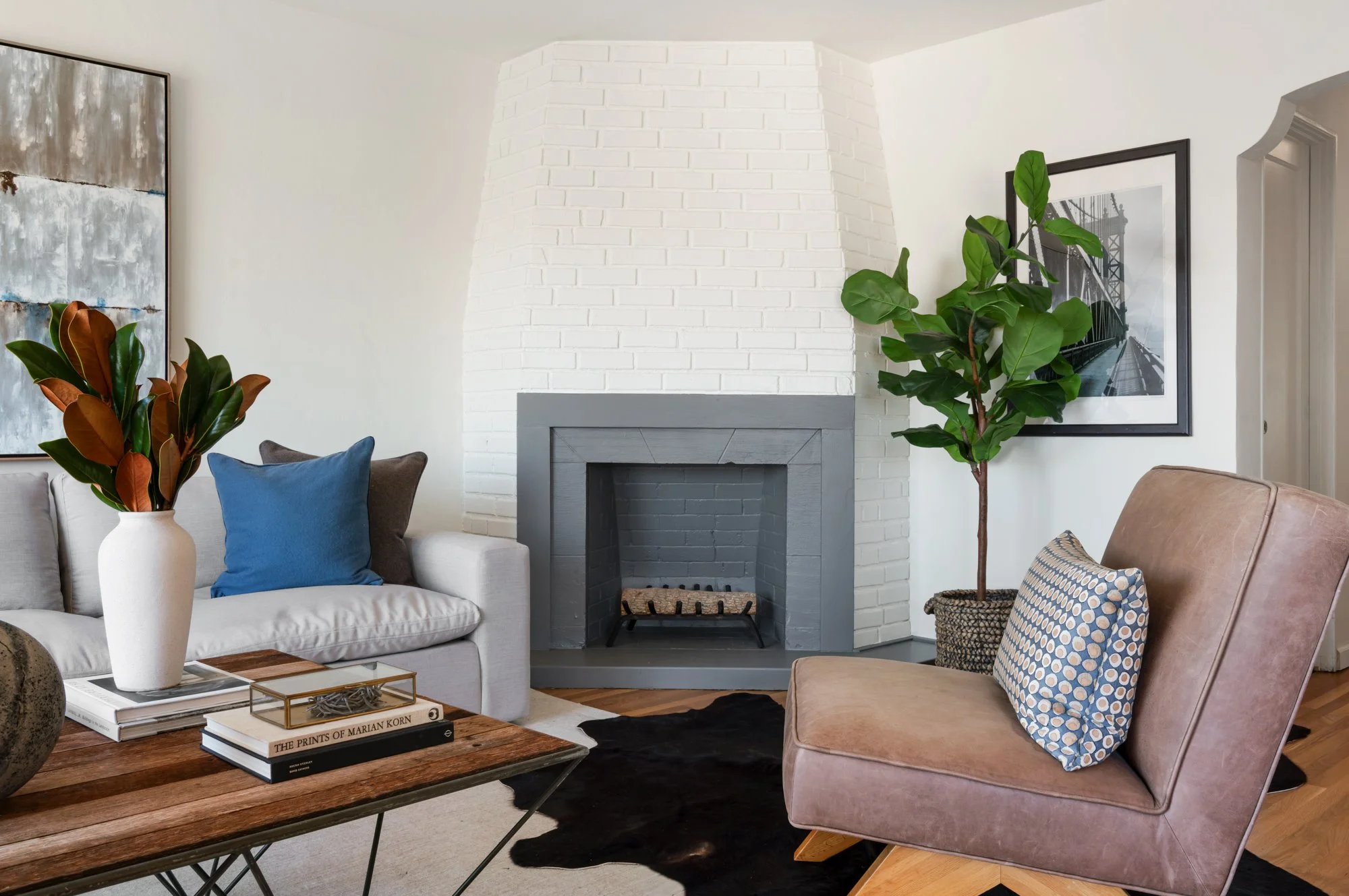 Modern living room with a white sofa, blue and gray pillows, a wooden coffee table with books and decorative items, a black and white photograph of a bridge on the wall, a large potted plant, and a stylish fireplace with white brick and gray accents.