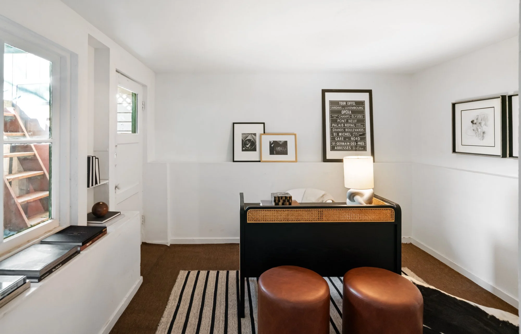 Interior of a small, minimalist living room with white walls, featuring framed black and white artwork, a black and wood side table with a white lamp, two brown leather stools, a window with books on the sill, and a striped rug.